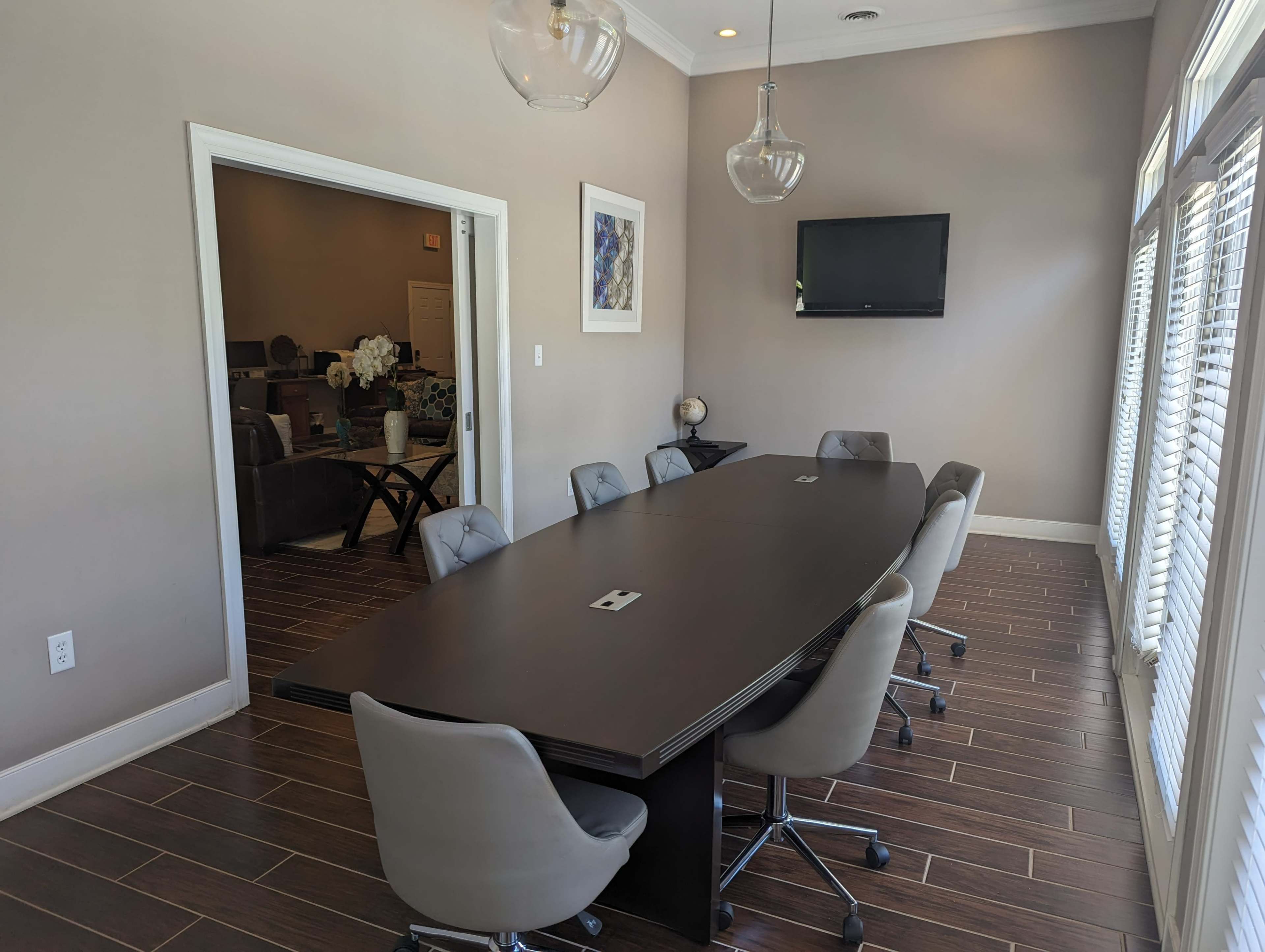 A modern conference room features a long black table surrounded by gray chairs, with a television mounted on the wall and windows allowing natural light.