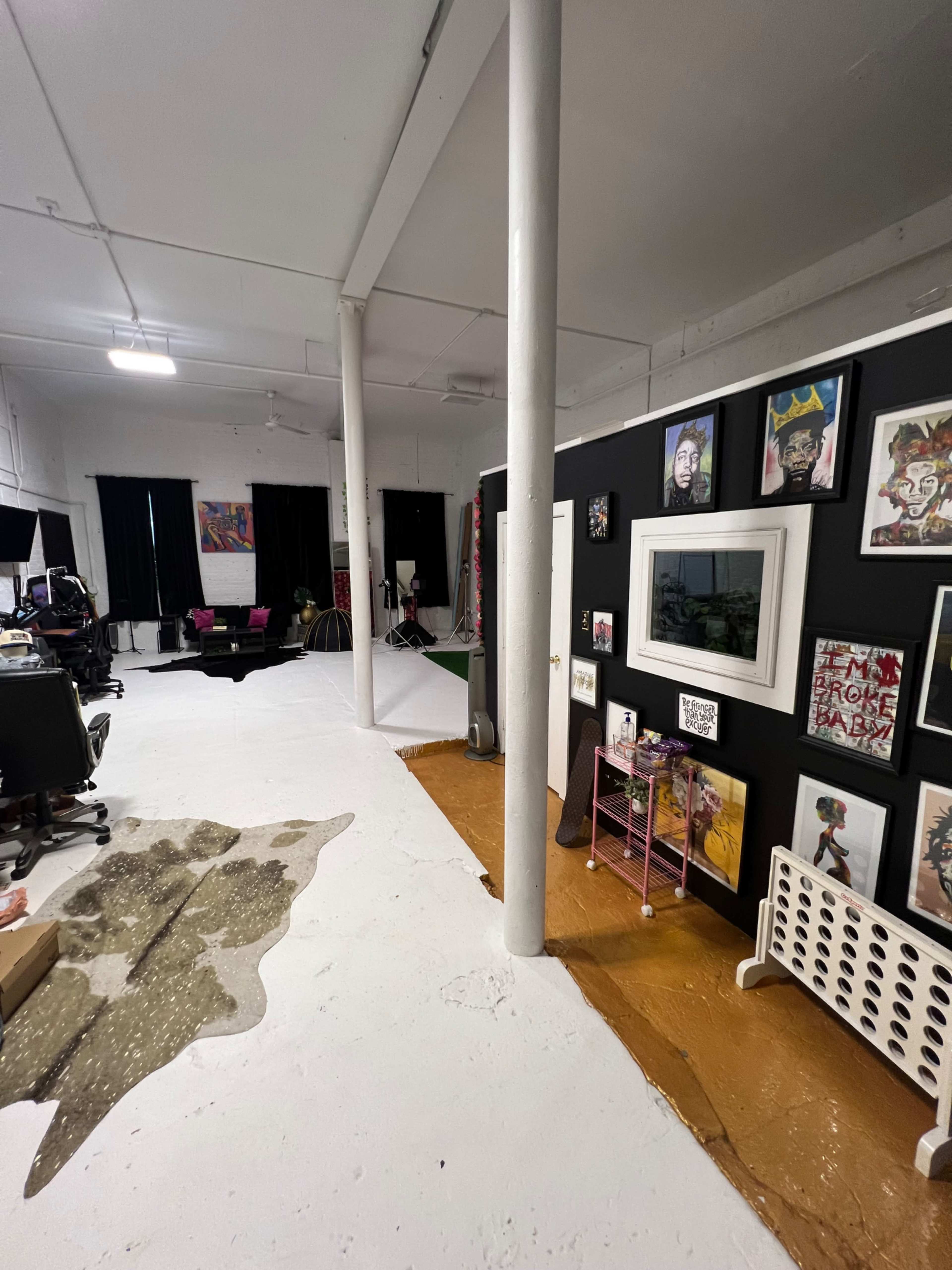 An open workspace with black walls adorned with framed art, a carpeted area, and scattered furniture in a well-lit room.