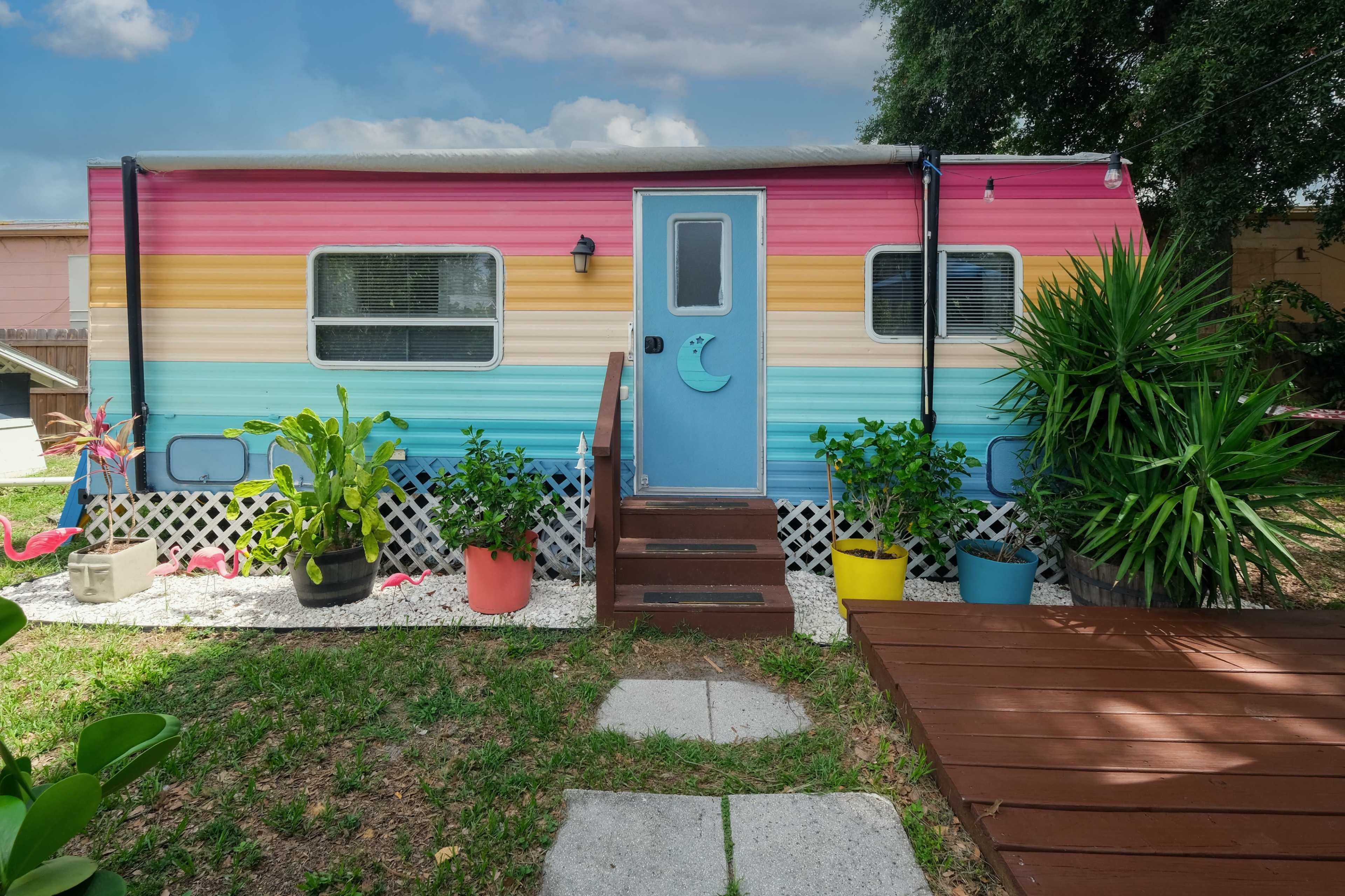 The image shows a colorful striped trailer with a blue door surrounded by potted plants and a wooden deck in a grassy area.