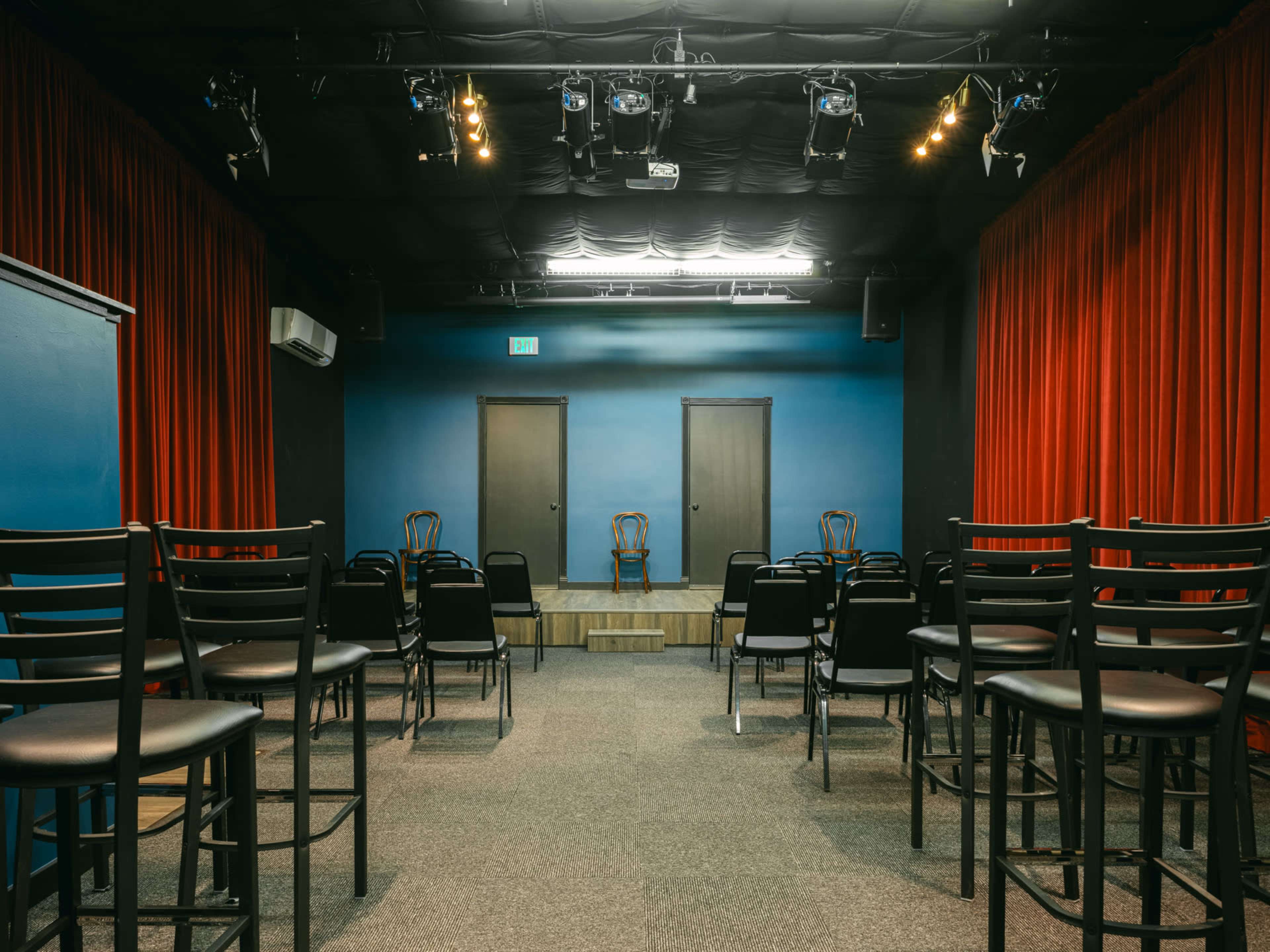 A small theater with black bar stools arranged in rows facing a raised wooden stage, flanked by two closed doors and red curtains.