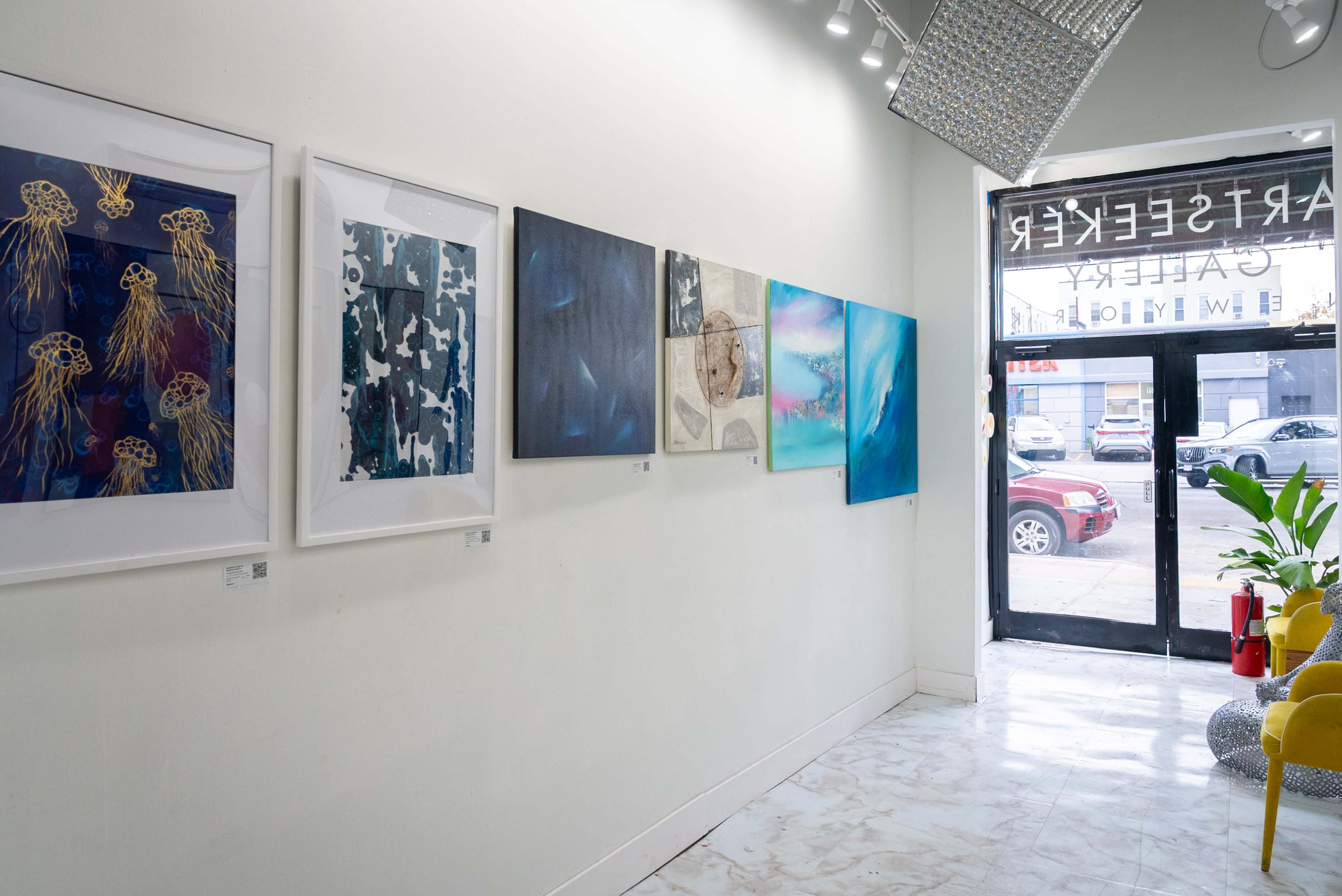The image shows an art gallery with a series of framed paintings displayed on the wall, featuring various abstract themes, while natural light streams in through large front windows.