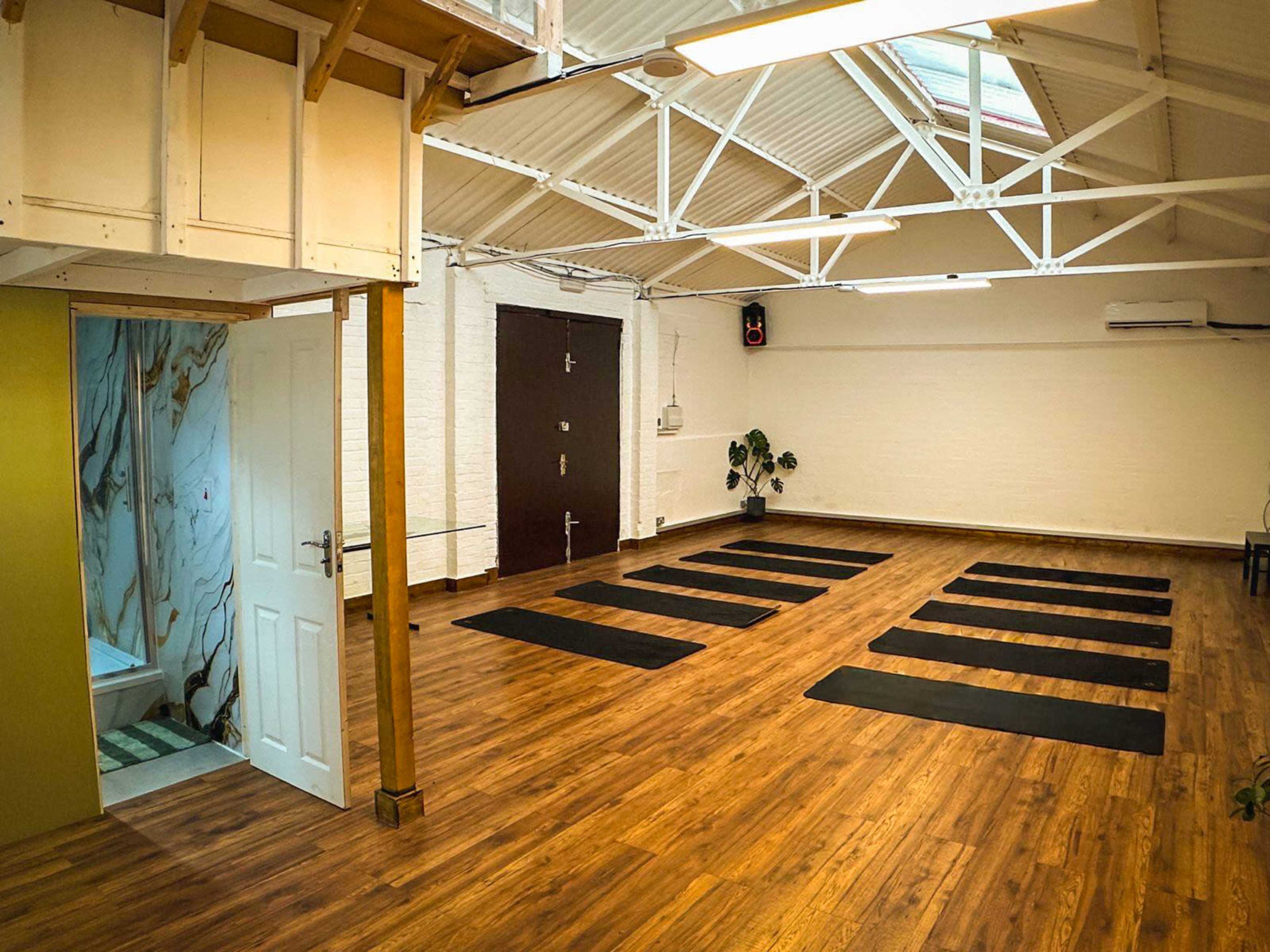 The image shows an empty yoga studio with wooden flooring, several yoga mats arranged in parallel, and a door leading to a restroom.