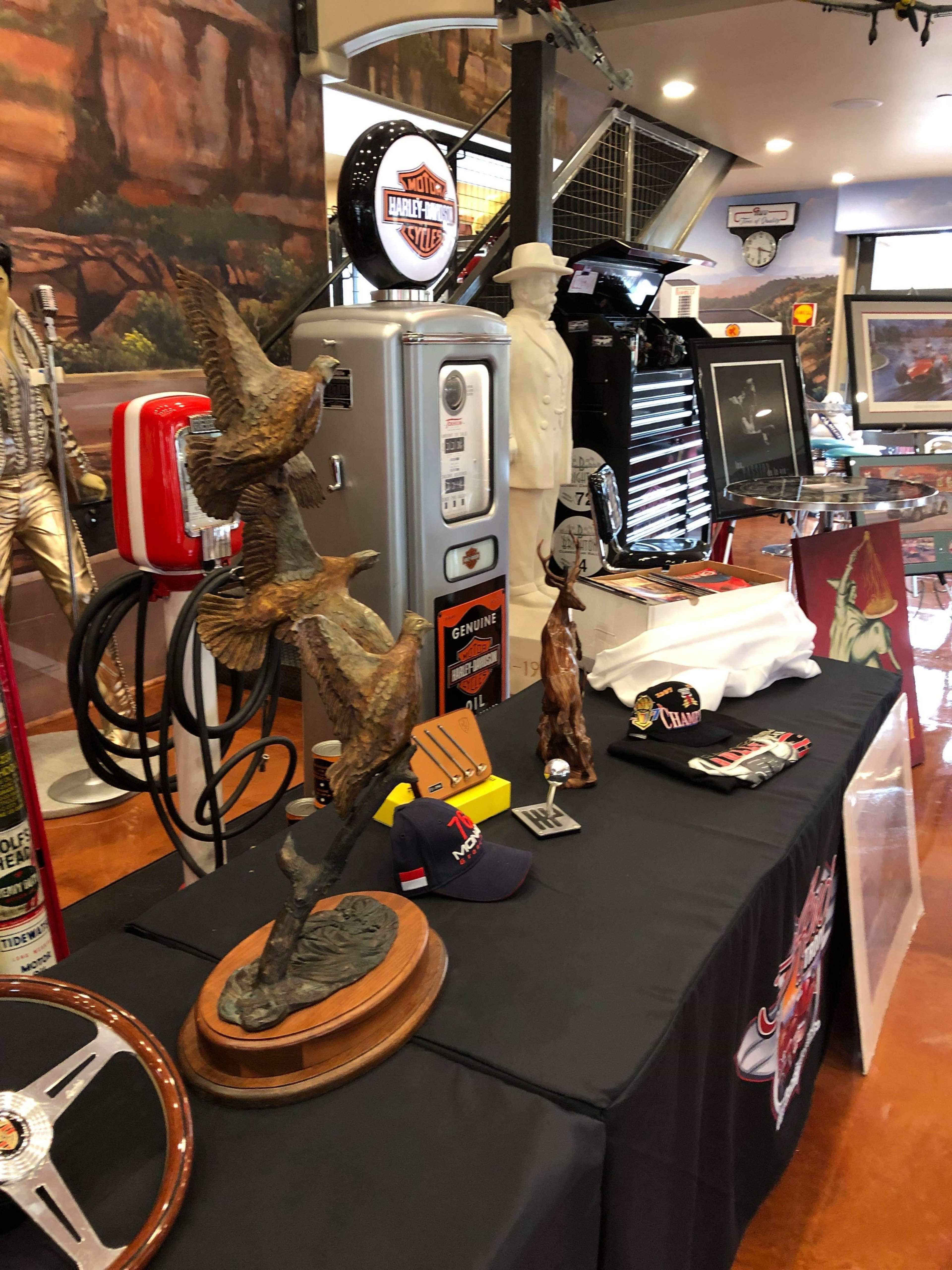 A display table with various items, including a bronze sculpture of birds, automotive memorabilia, caps, and a toolbox, set in a themed interior space.