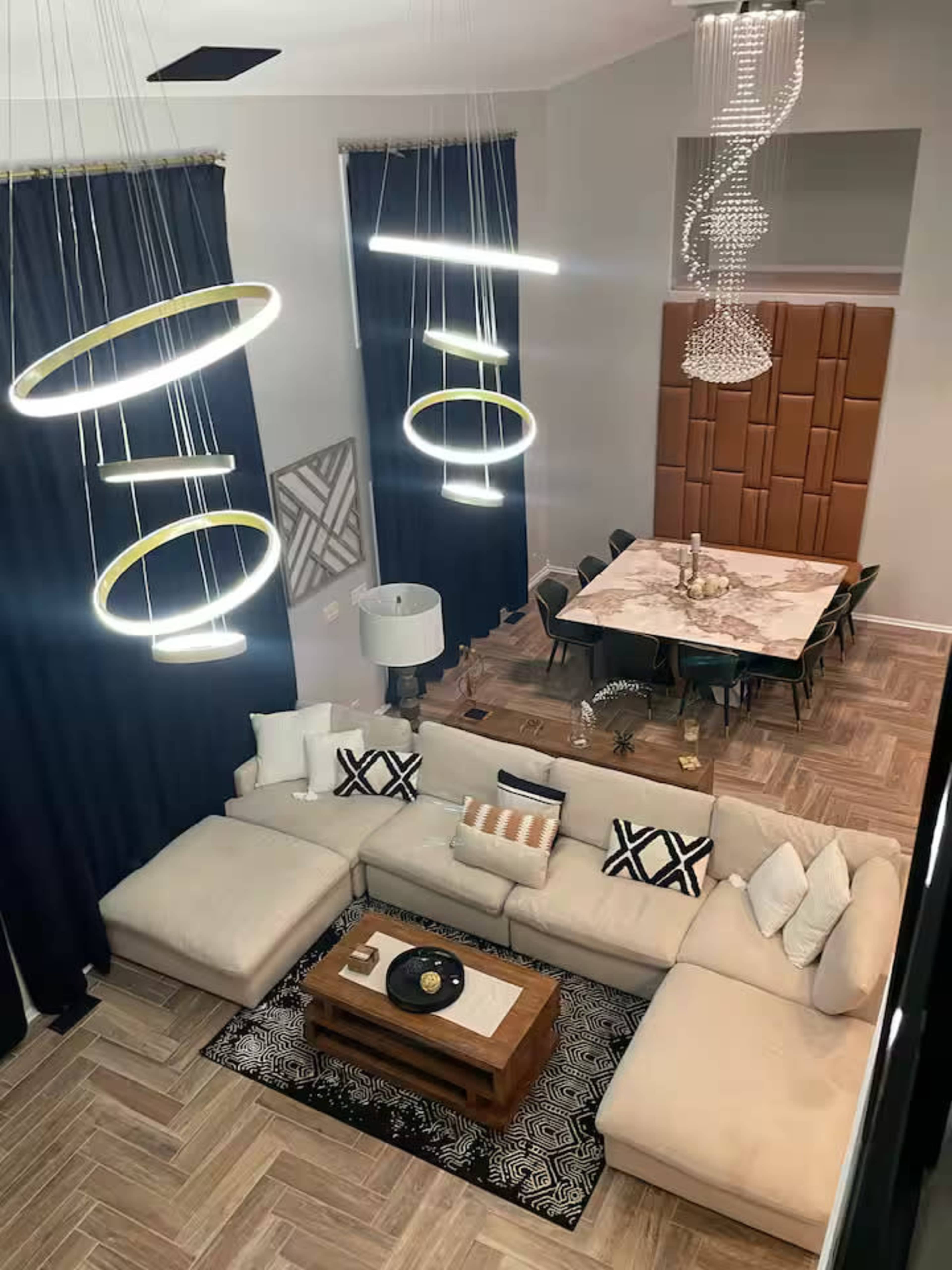 The image shows a spacious living area featuring a large sectional sofa, a round coffee table, modern pendant lights, and a dining table set nearby.