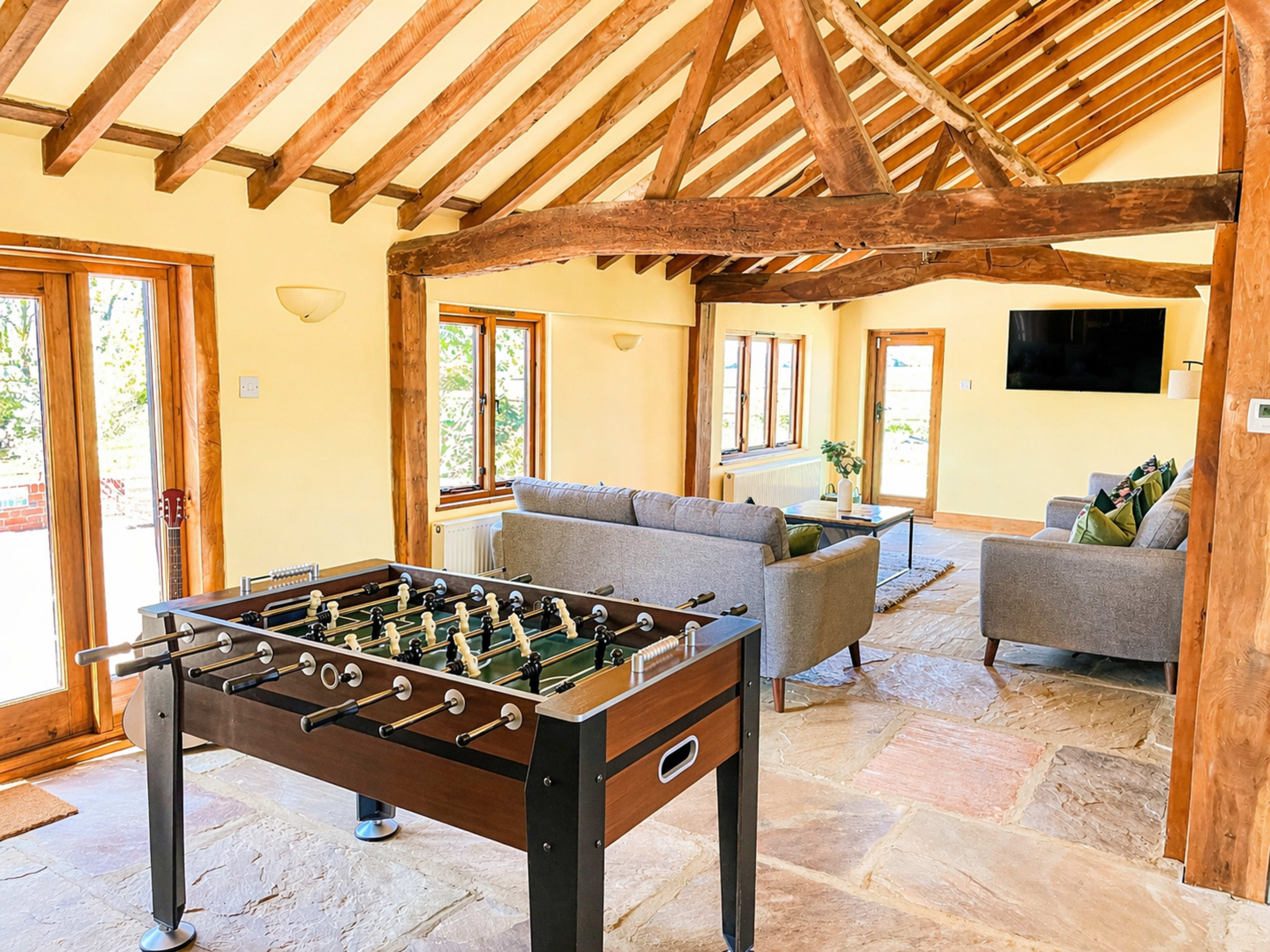 The image shows a spacious room with exposed wooden beams, a foosball table in the foreground, and a seating area with gray sofas and large windows.