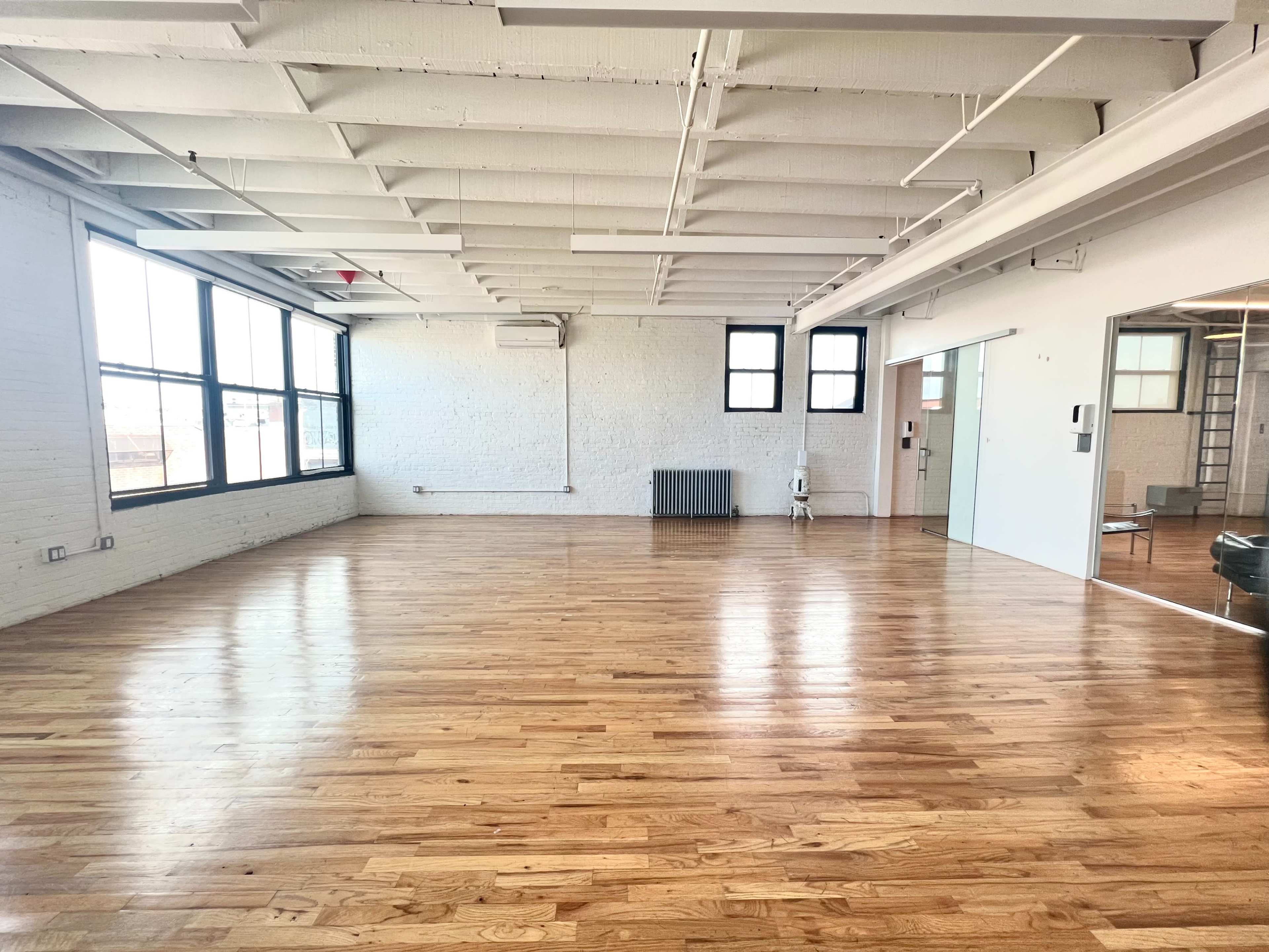 The image shows an empty room with hardwood floors, large windows, exposed brick walls, and a sliding glass door.