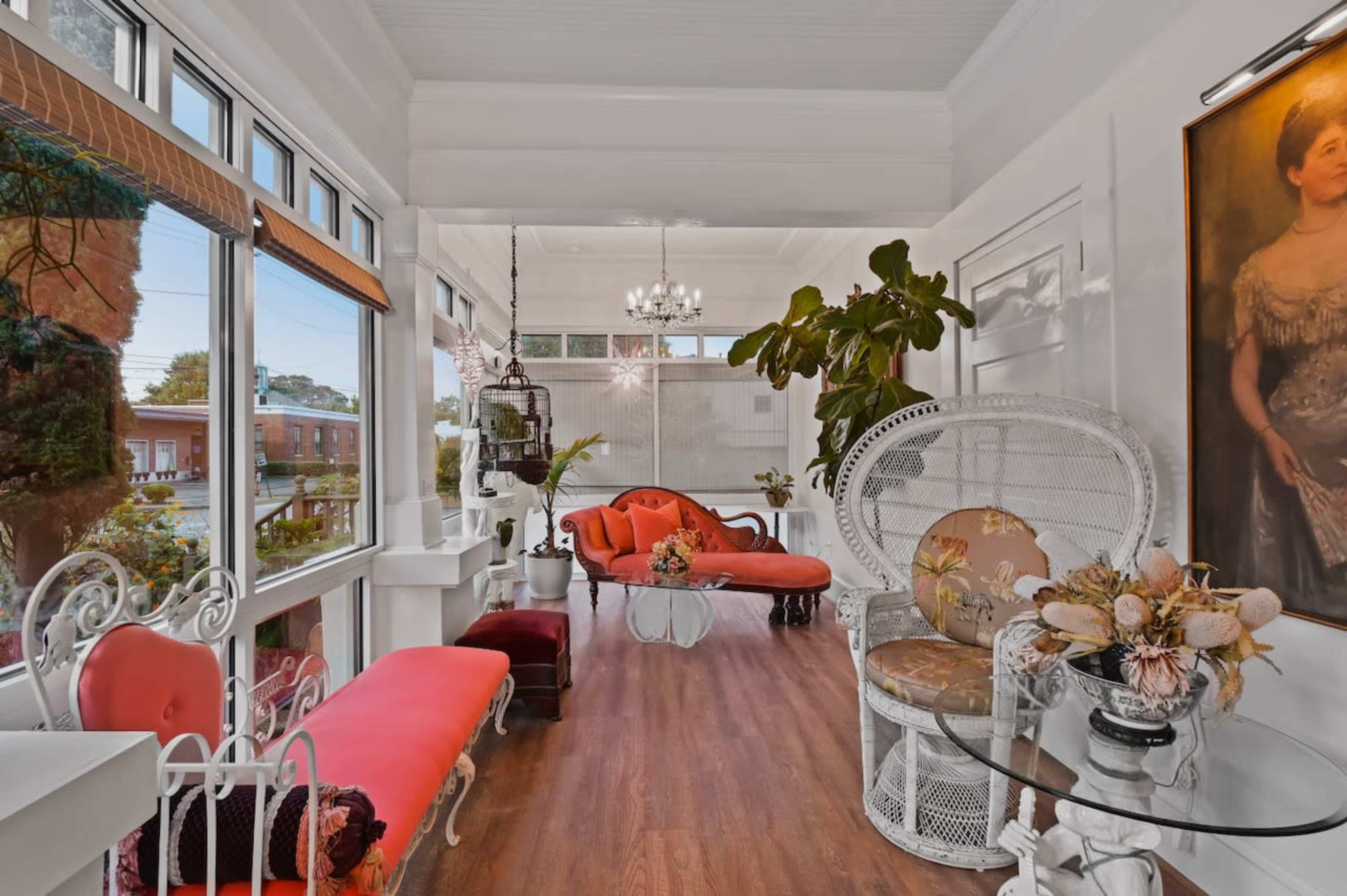A sunlit room featuring red seating, a glass table, and decorative plants, with large windows allowing natural light.