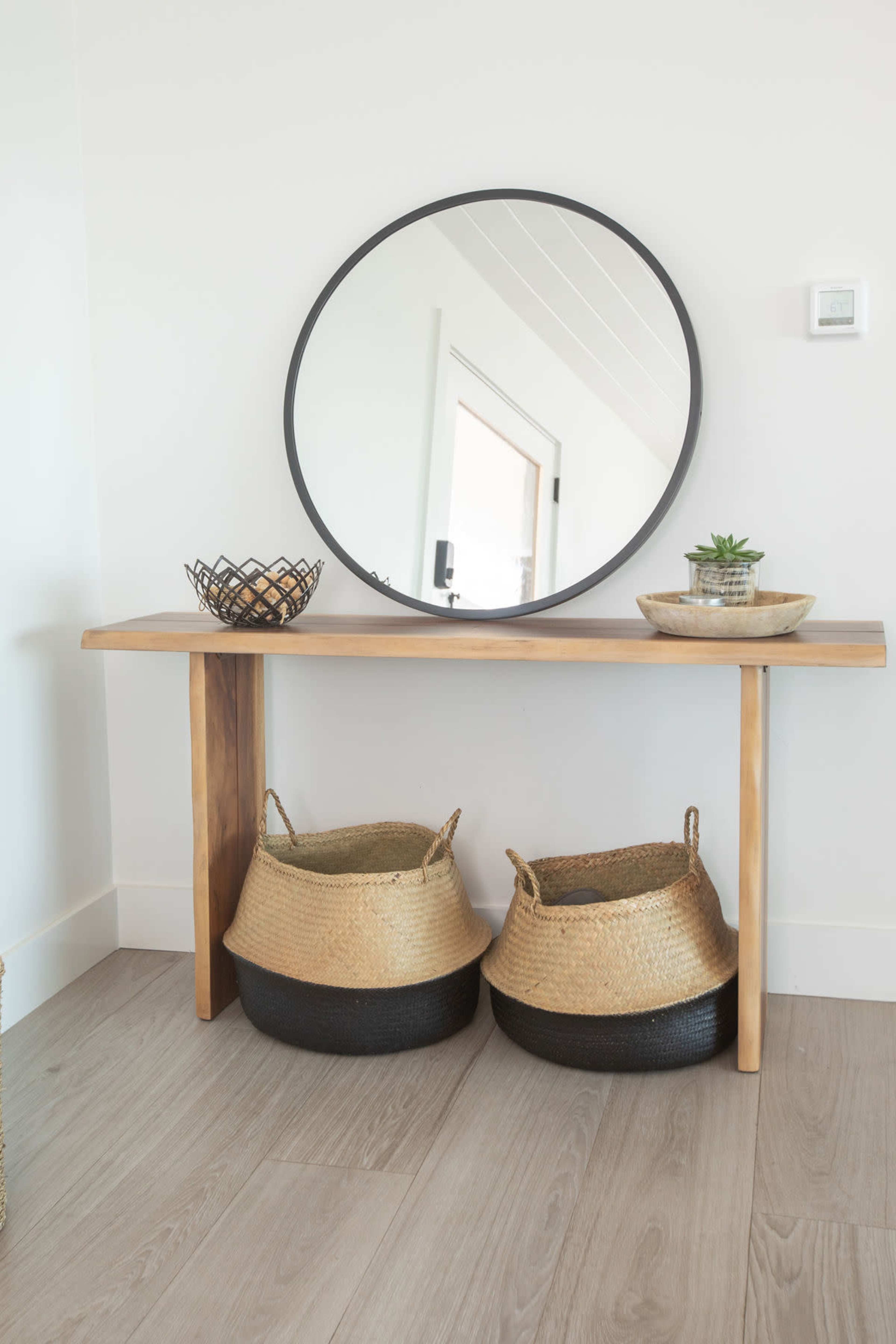 A wooden console table with a round mirror above it, flanked by two woven baskets and decorative bowls.
