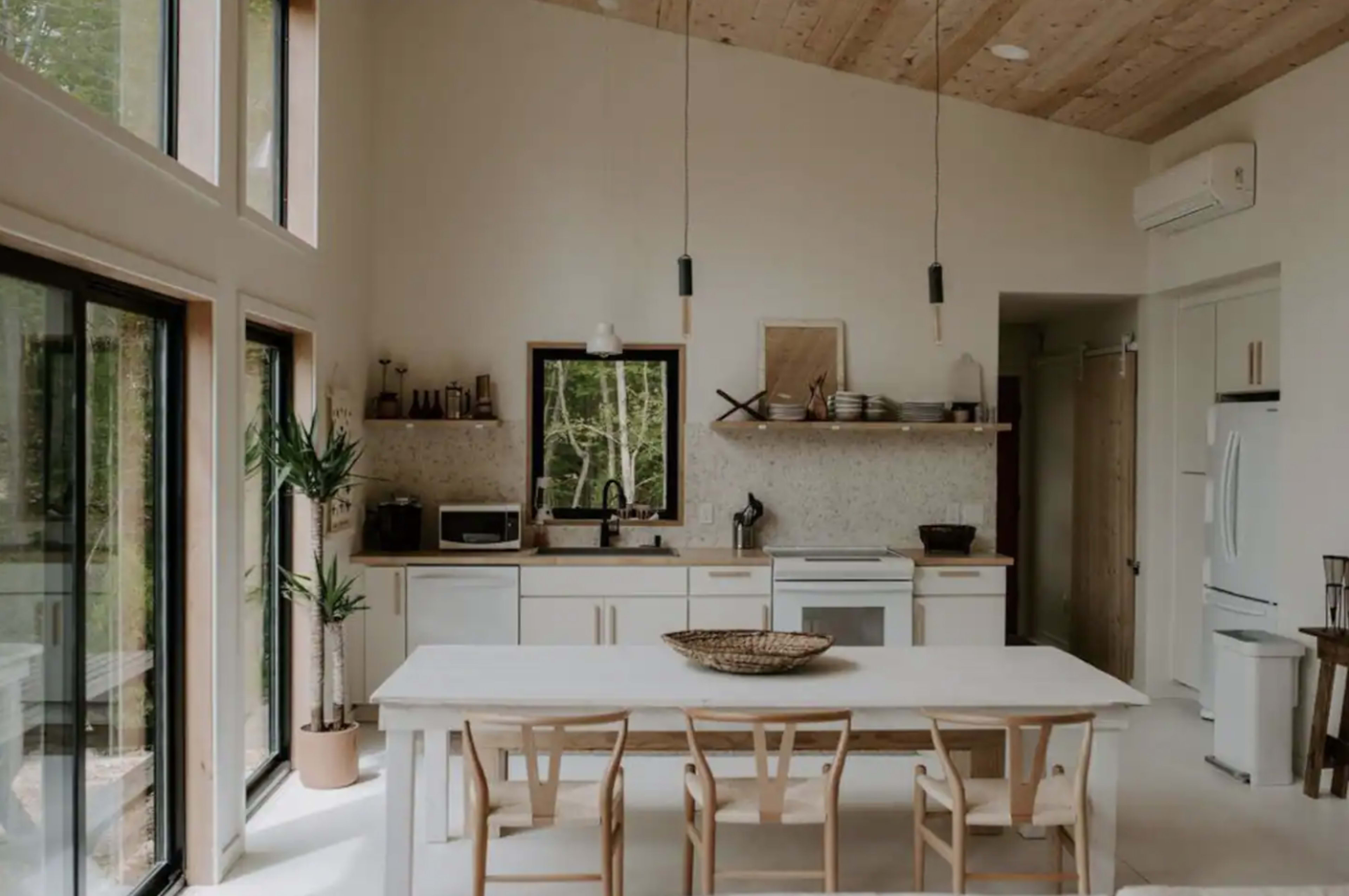 A modern, minimalist kitchen features a large white table surrounded by wooden chairs, with windowed walls allowing natural light and views of greenery outside.