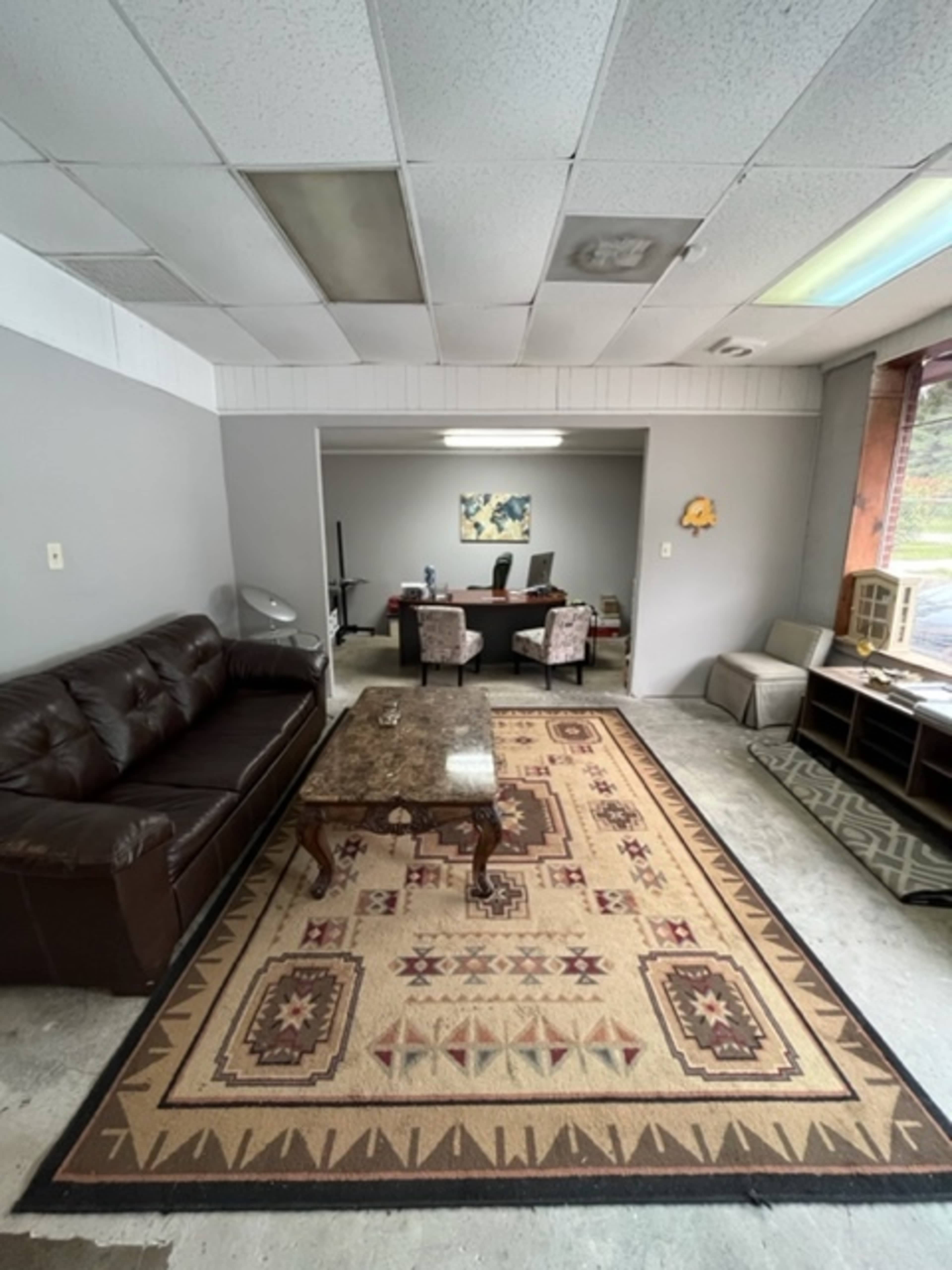 The image shows a small office space featuring a brown leather sofa, a patterned area rug, and a view into a separate meeting area with a desk and chairs.