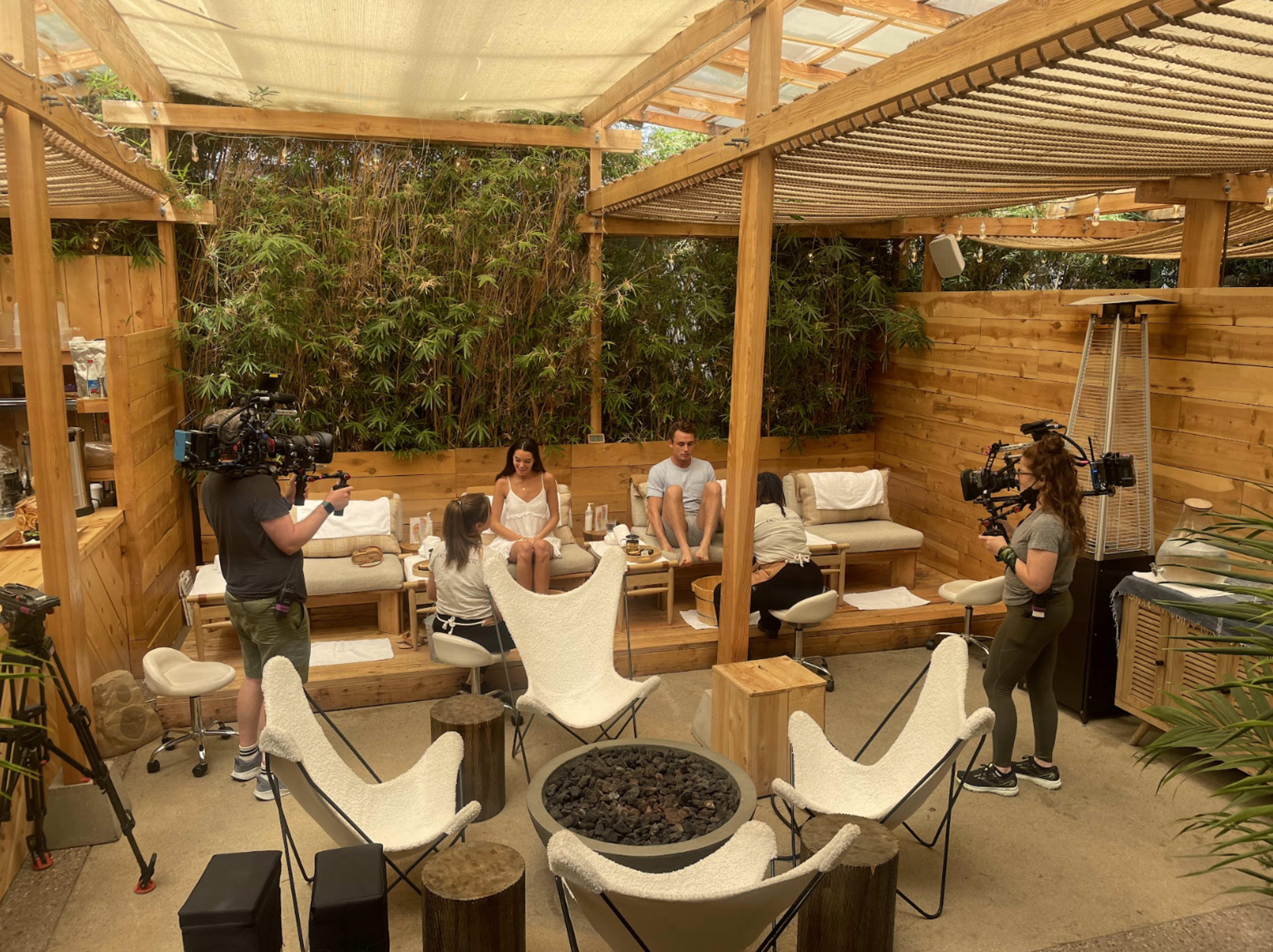 A wooden outdoor space with a filming crew capturing a scene involving two people seated on a bench surrounded by greenery.