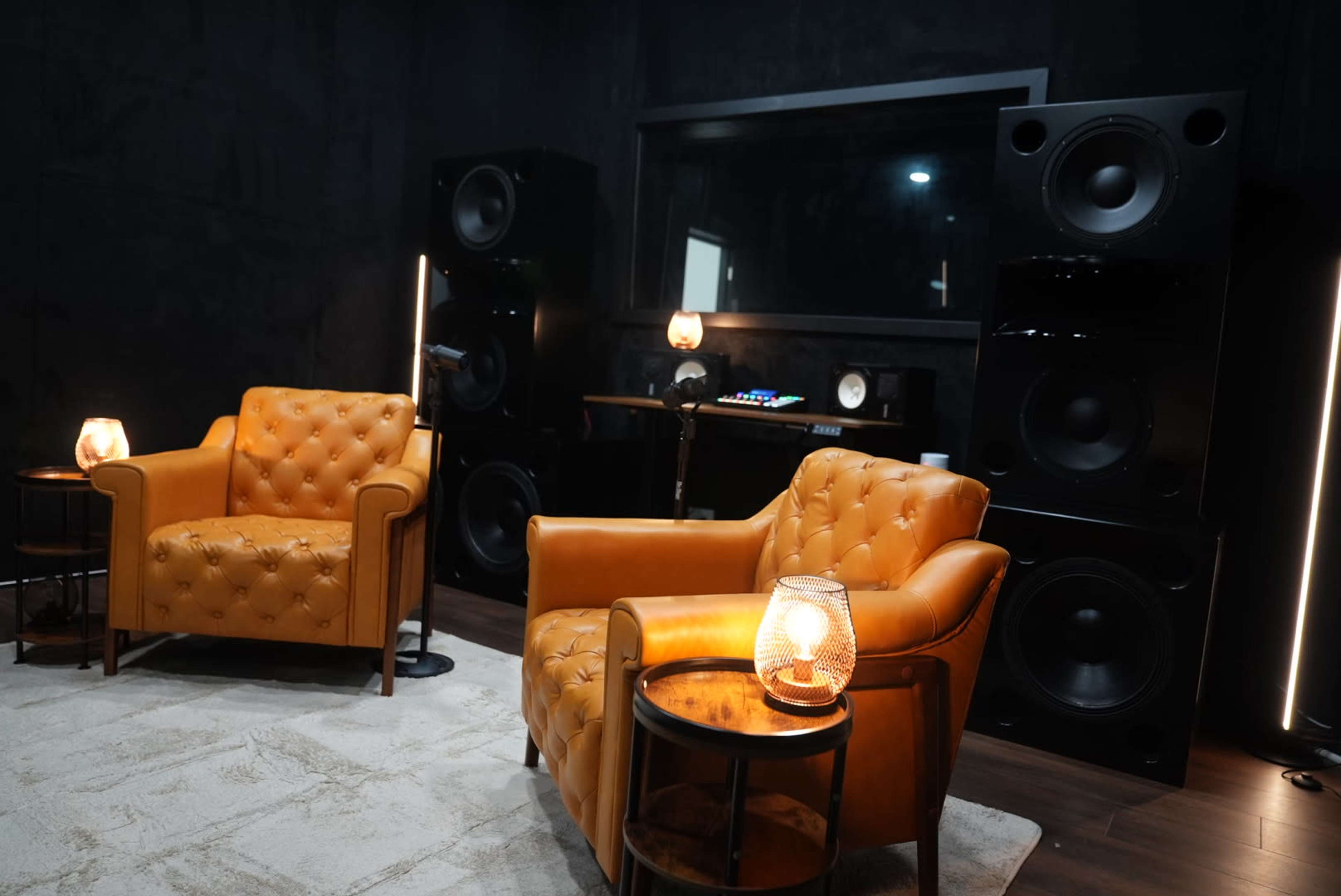 The image shows a small, stylish music studio featuring two brown leather armchairs positioned next to a table with a lamp, set against a backdrop of large speakers and a sound mixing station.