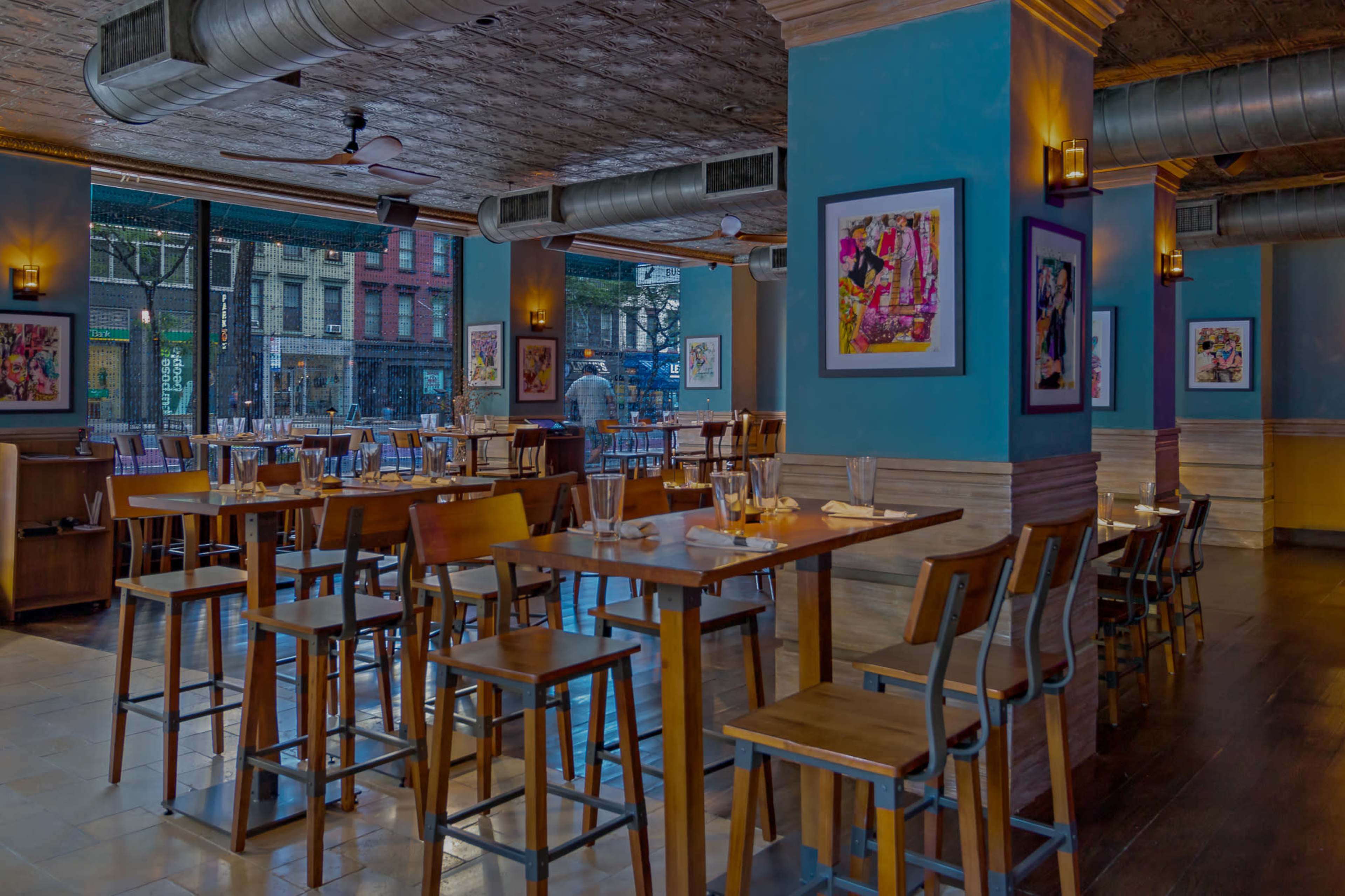 The image shows a spacious restaurant with wooden tables and chairs, large windows, and colorful artwork on the walls.