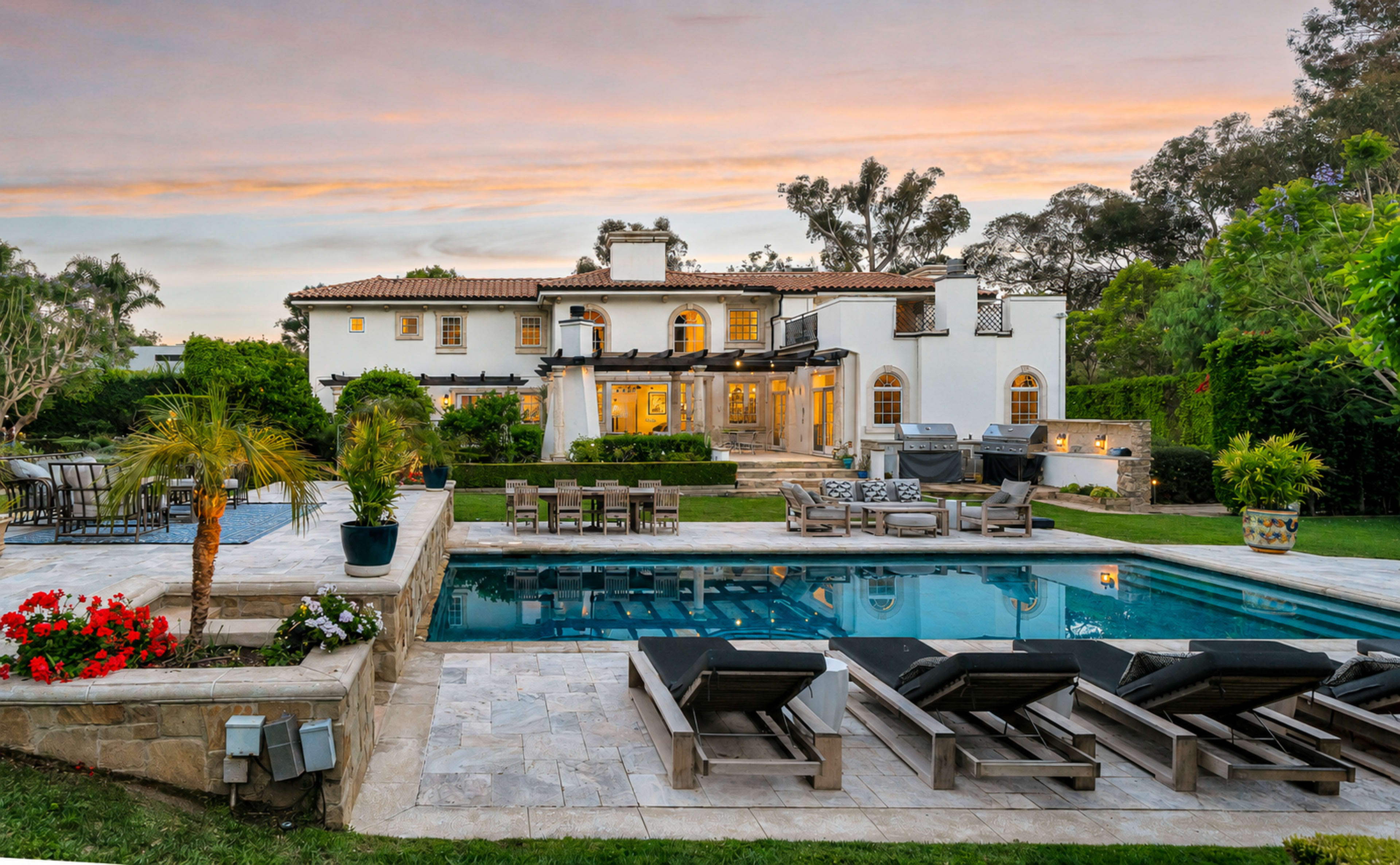 A luxurious two-story white villa with a large swimming pool and outdoor seating area is set against a colorful sunset.