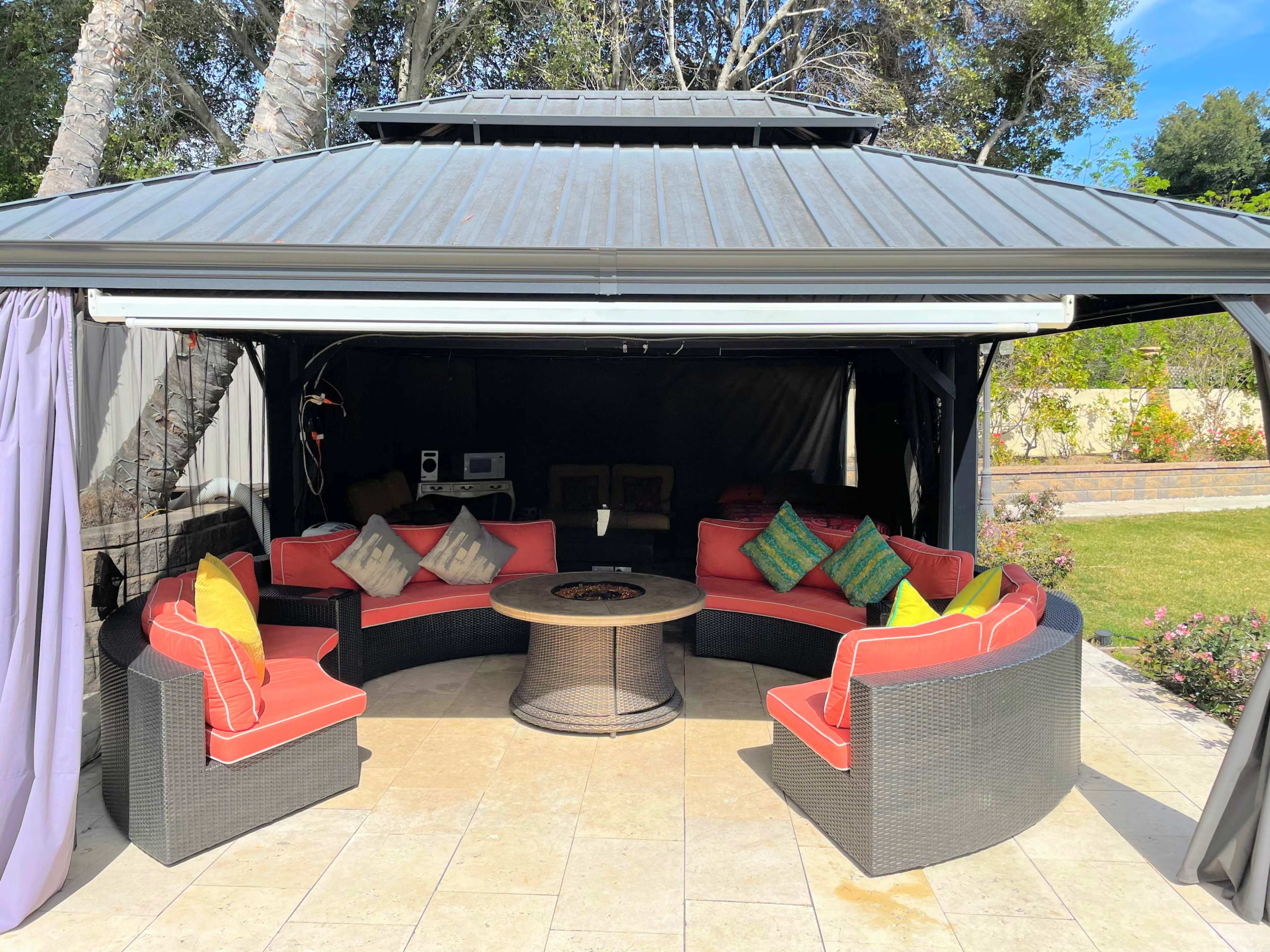 A circular seating area with red cushions surrounds a central table under a gazebo with a metal roof and gray walls.