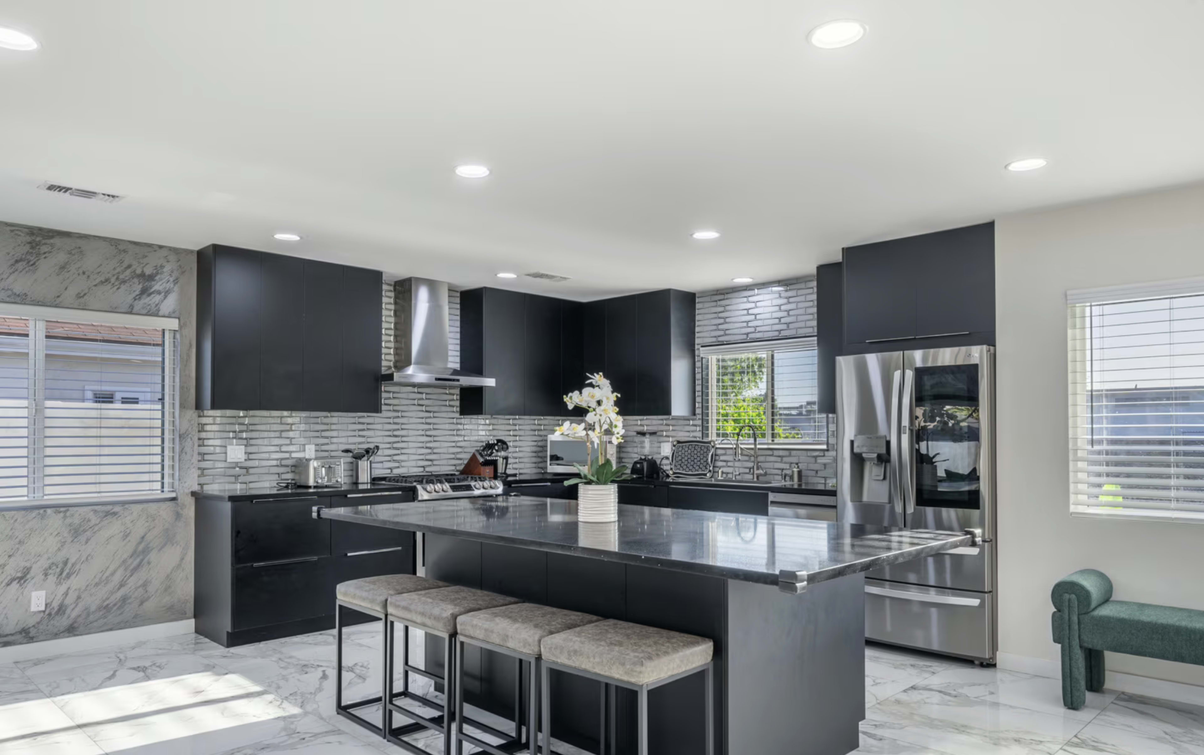 The image shows a modern kitchen with dark cabinetry, a large marble island, and stainless steel appliances, complemented by white walls and a marble floor.