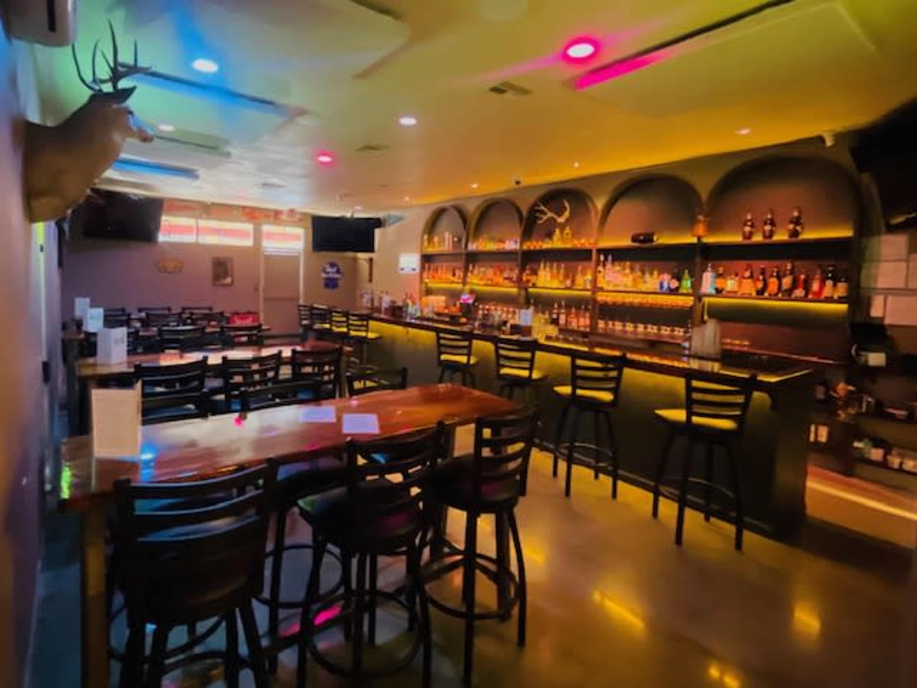 The image depicts a modern bar interior featuring a well-lit counter with shelves of liquor bottles and several tables with chairs arranged around the space.
