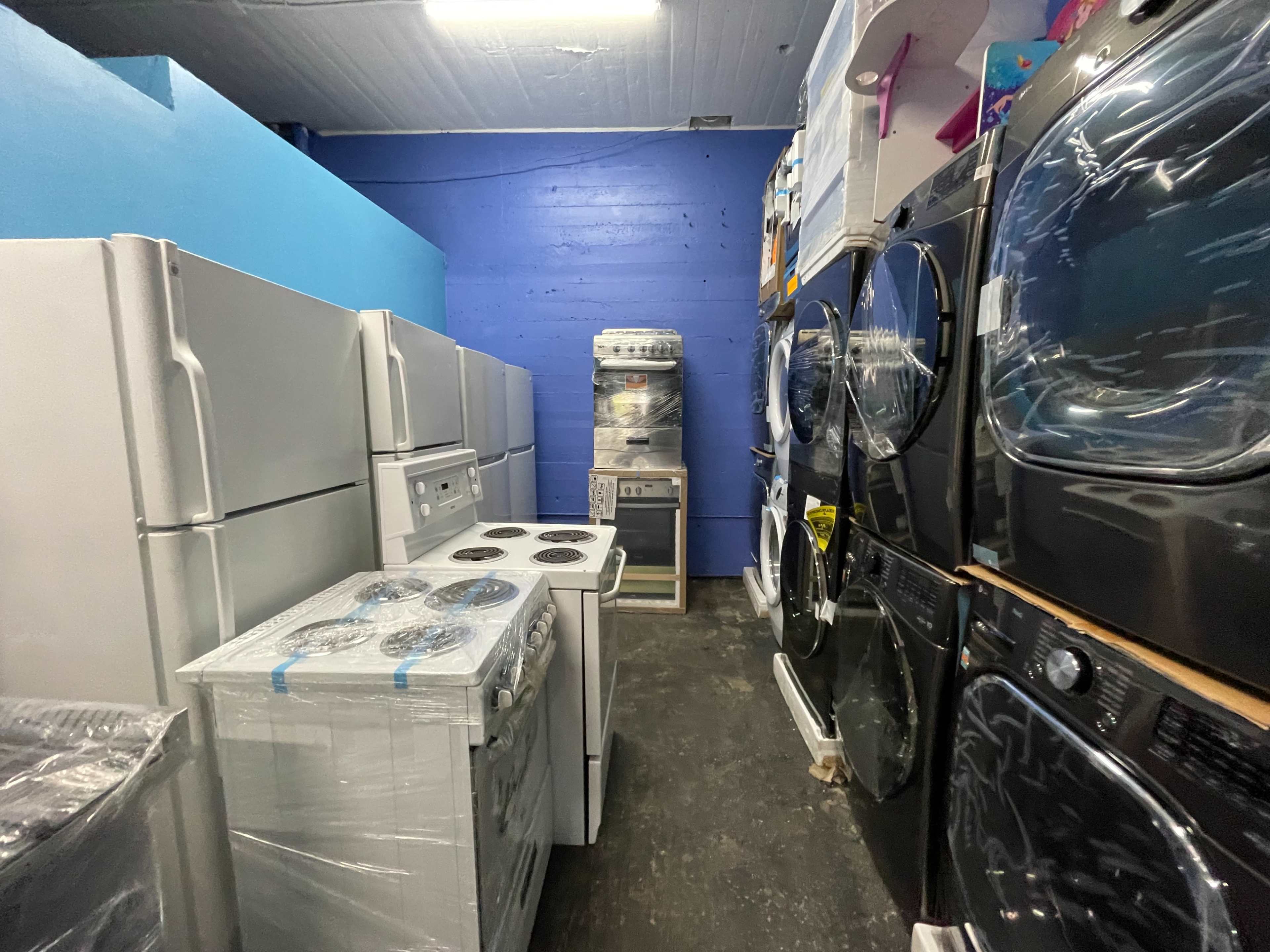 The image shows a narrow aisle lined with various kitchen appliances, including white refrigerators, a stove, and black washer-dryer units, all displayed in a blue-walled room.