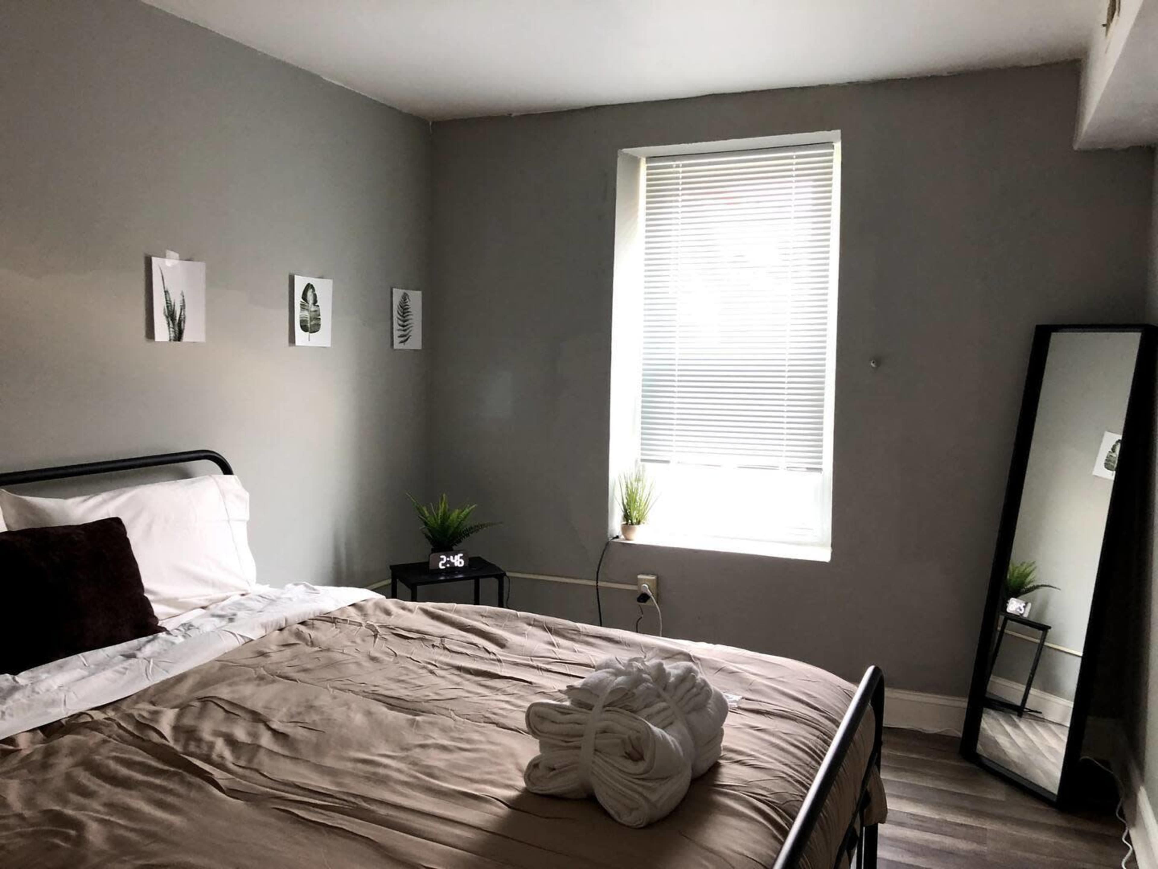 A simple bedroom features a bed with beige linens, a large mirror, a window with blinds, and plants on either side.