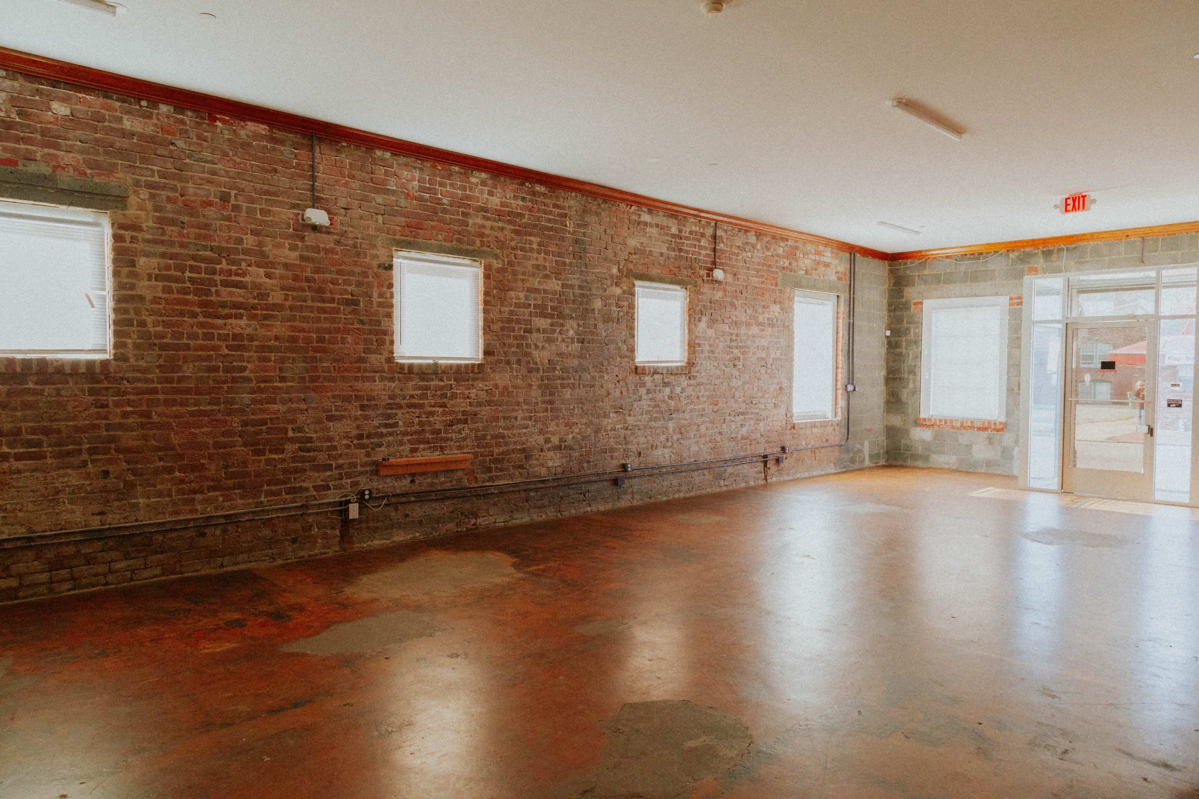 The image shows an empty room with exposed brick walls and large windows, featuring a concrete floor and an entrance door.