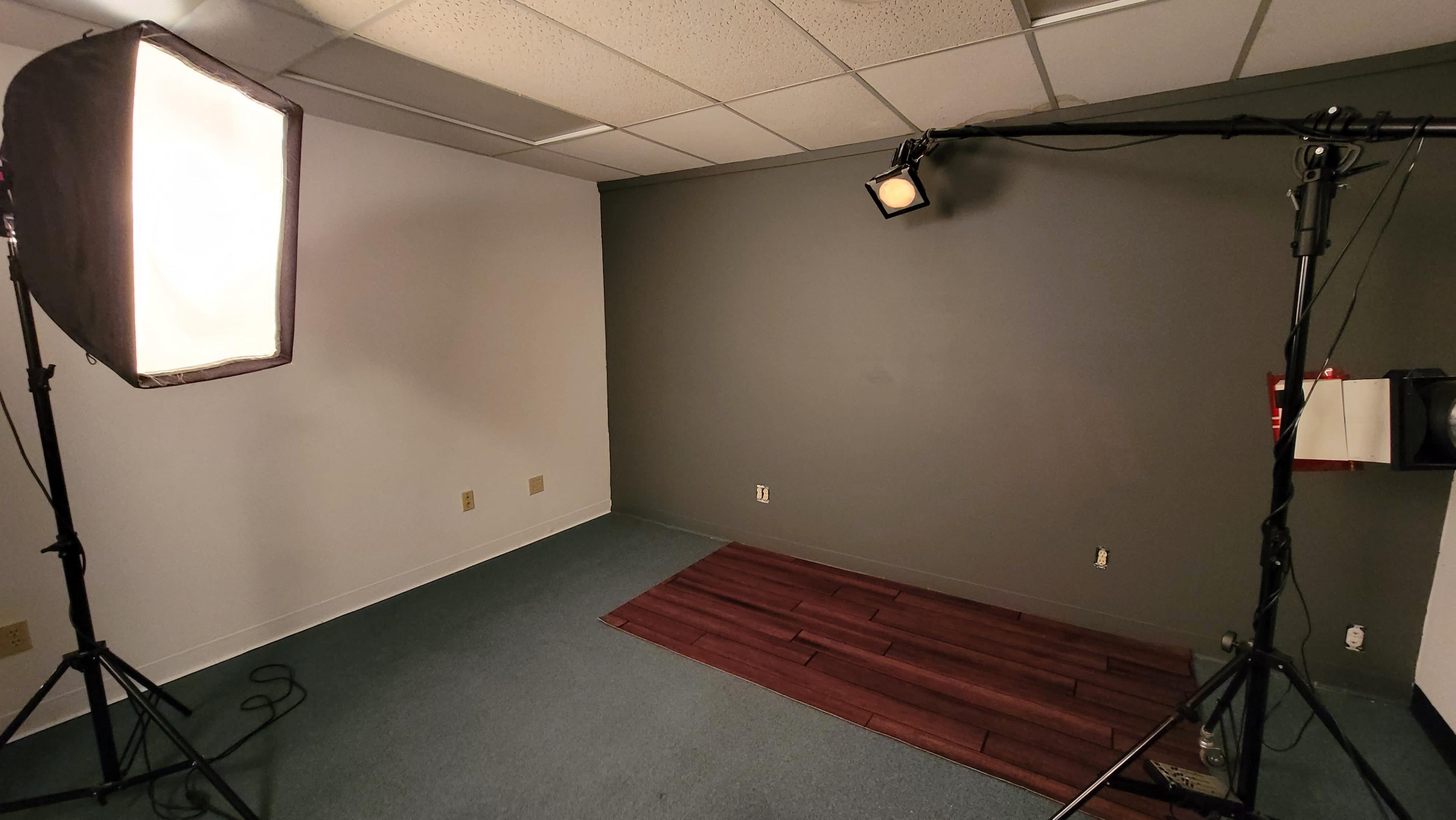 The image shows a sparsely furnished studio room with a dark gray wall, a wooden floor section, and two lighting setups on tripods.