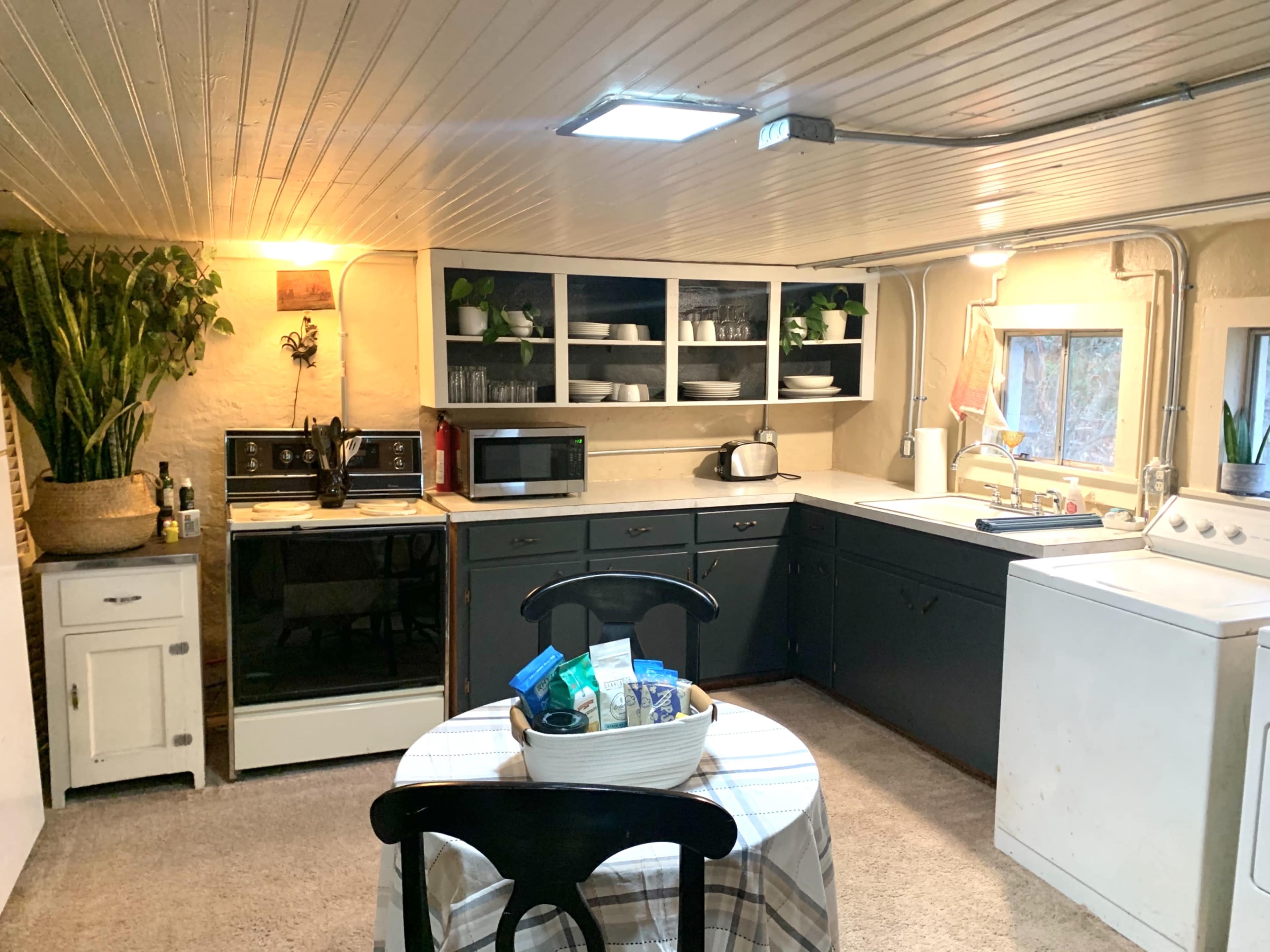 The image shows a cozy kitchen with white and dark cabinetry, appliances including a microwave and fridge, a round table set in the center, and a laundry area.
