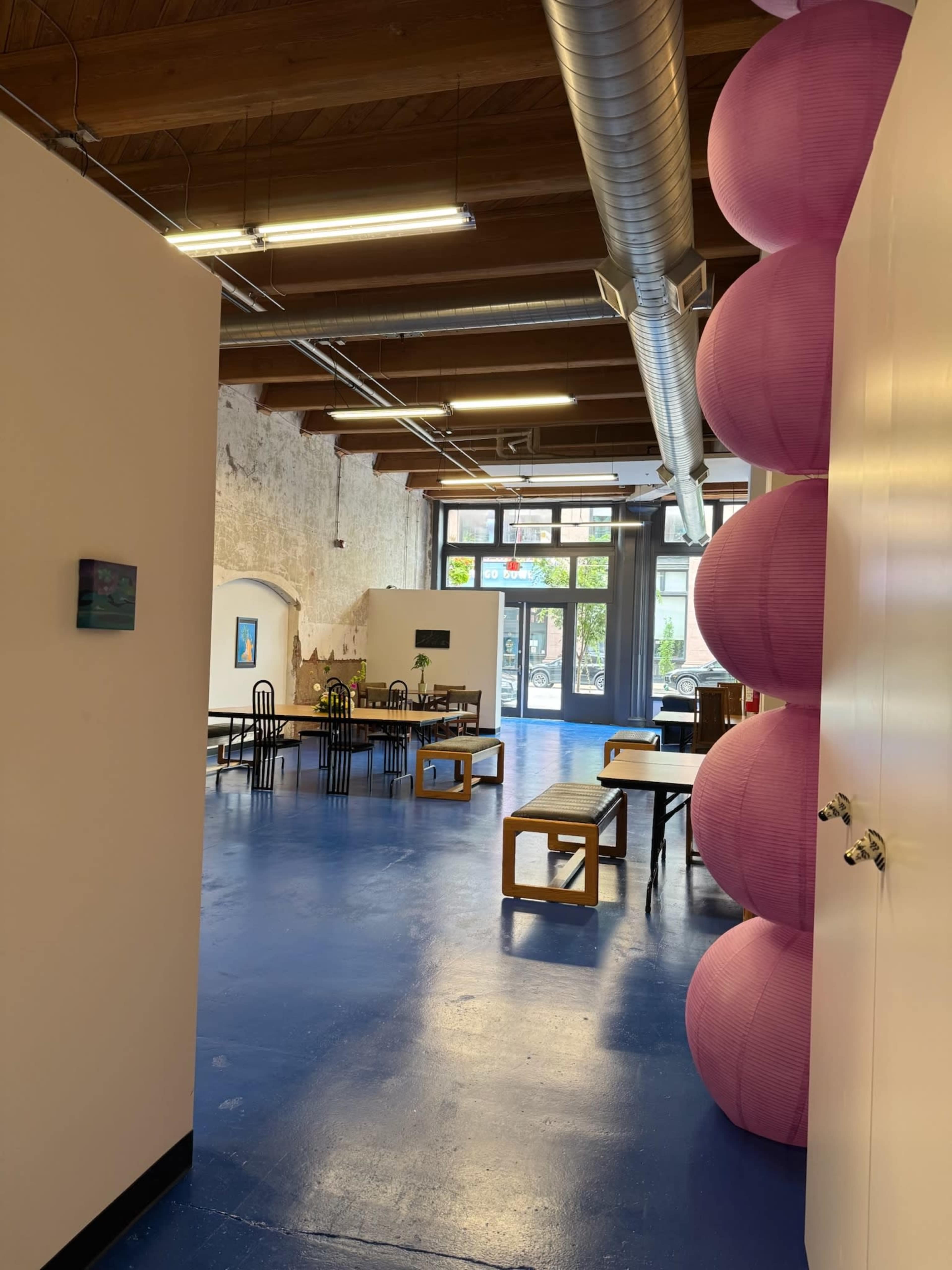 The image shows a spacious interior of a room with a blue floor, wooden beams, and large pink paper lanterns, leading toward tables and chairs in a well-lit area.