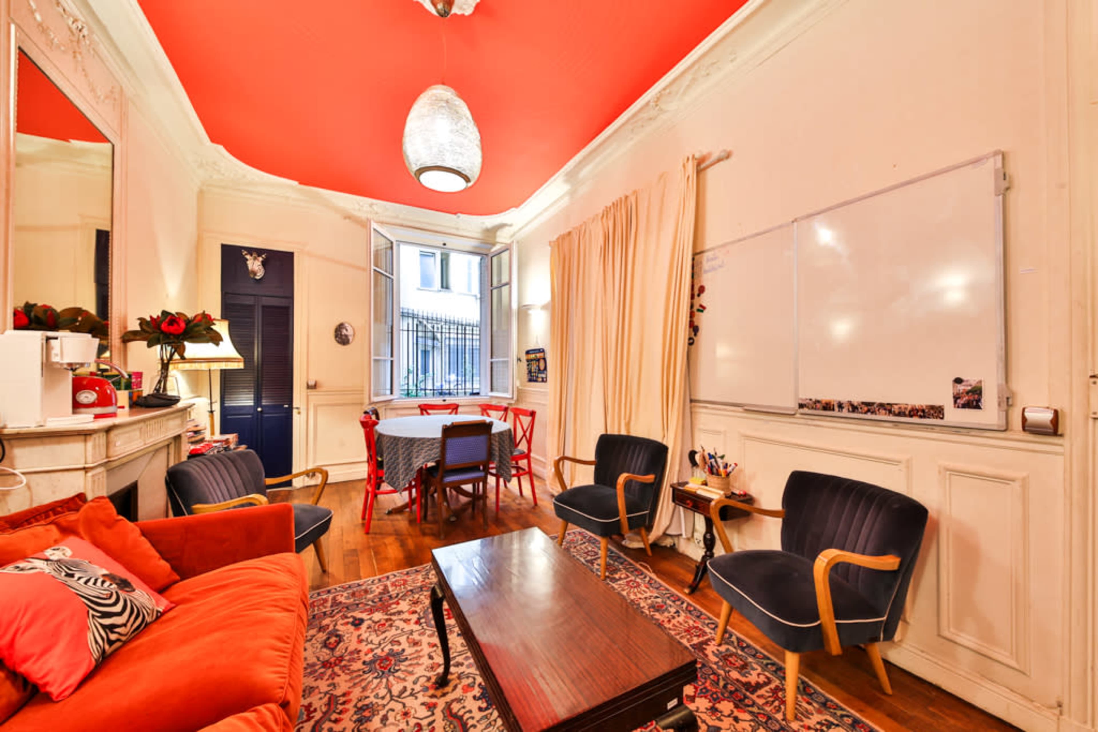 The image shows a well-lit room with a red ceiling, a mix of seating options, and a whiteboard mounted on the wall.