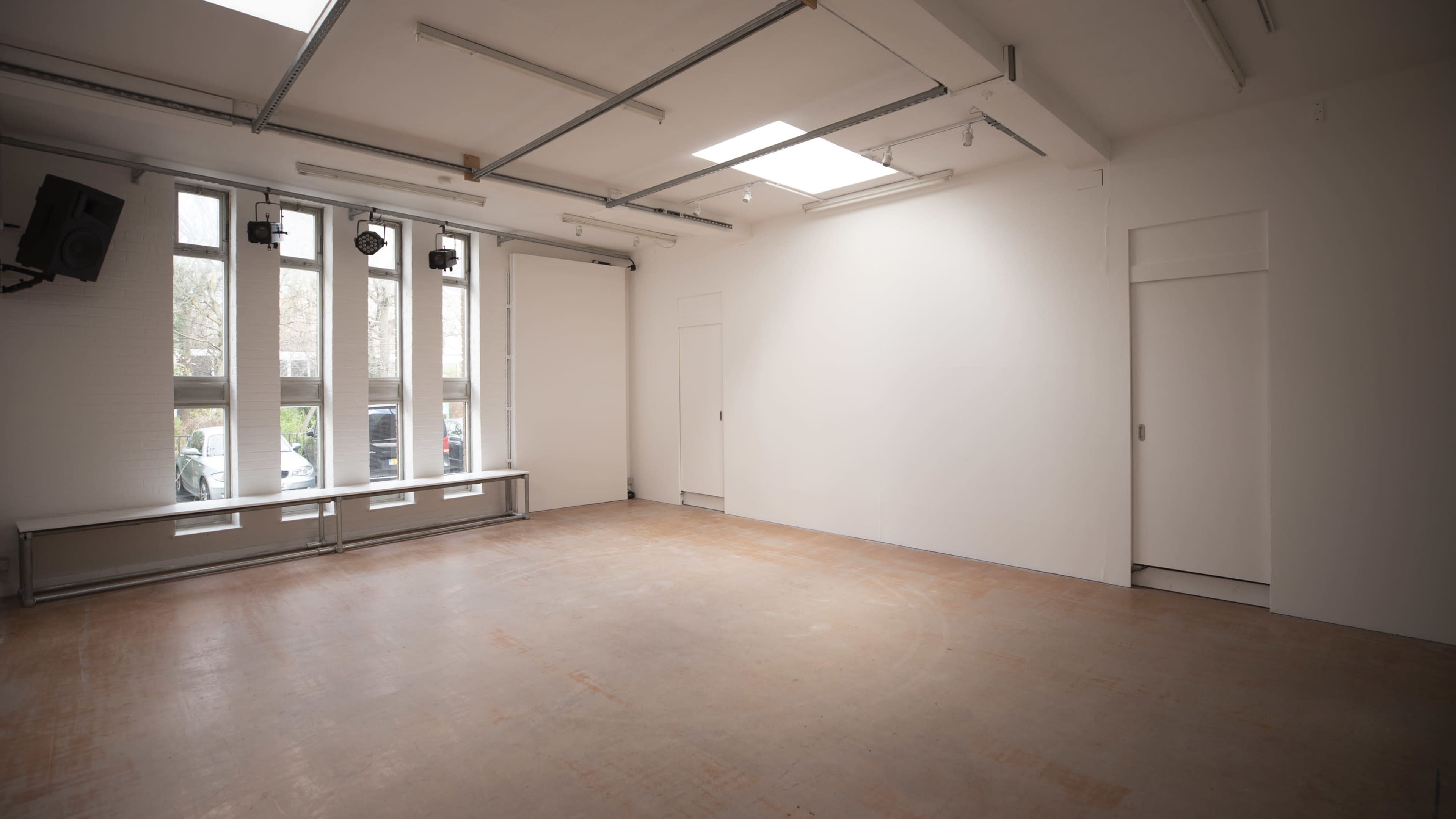 An empty room features large windows on one side, light-colored walls, and a wooden floor.