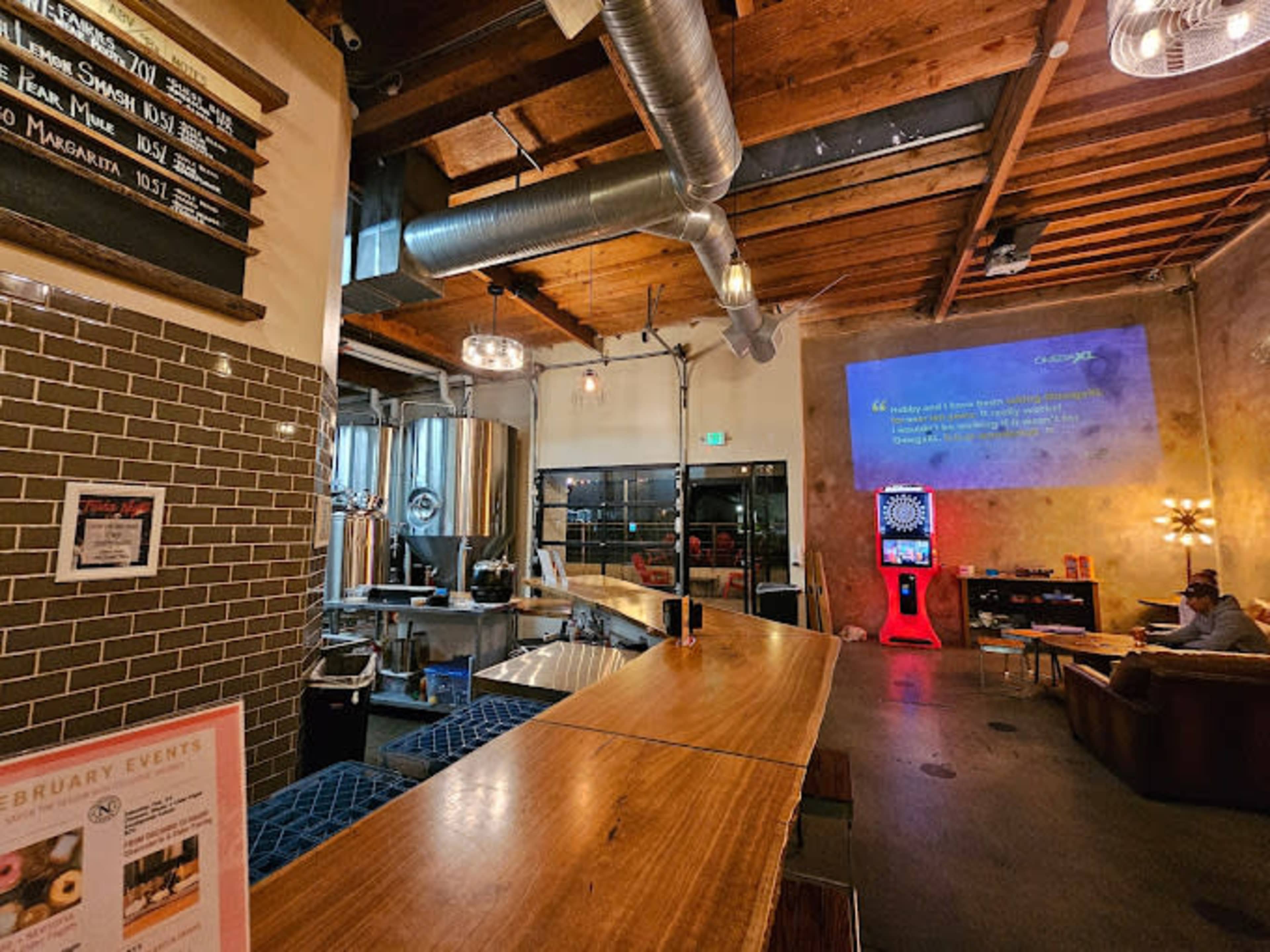 The image shows a modern bar area with a wooden countertop, brewing equipment in the background, and a projection screen displaying content on a wall.