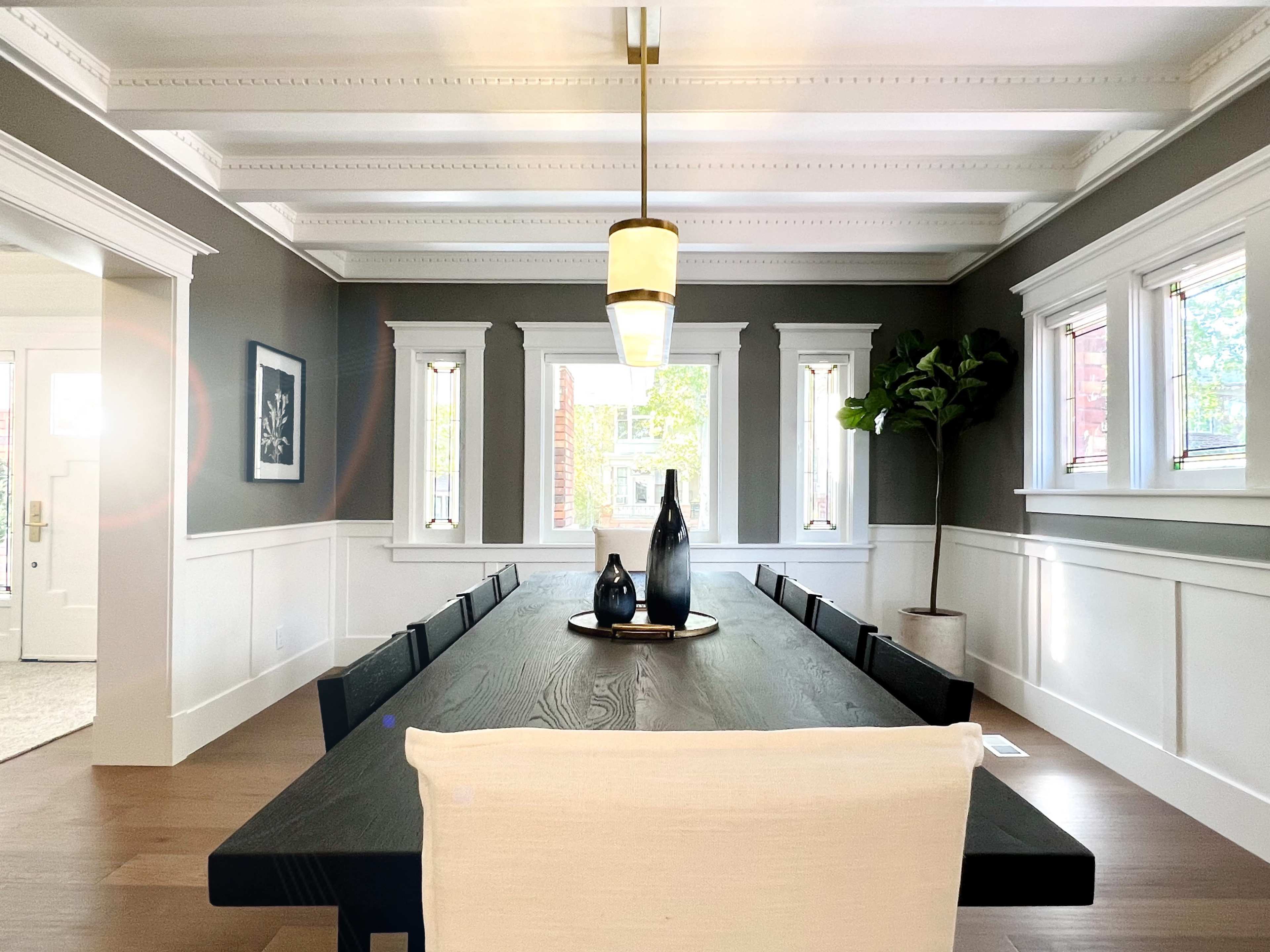 A long black dining table is set in a room with gray walls, white wainscoting, and large windows, featuring two decorative vases on the table and a potted plant in the corner.