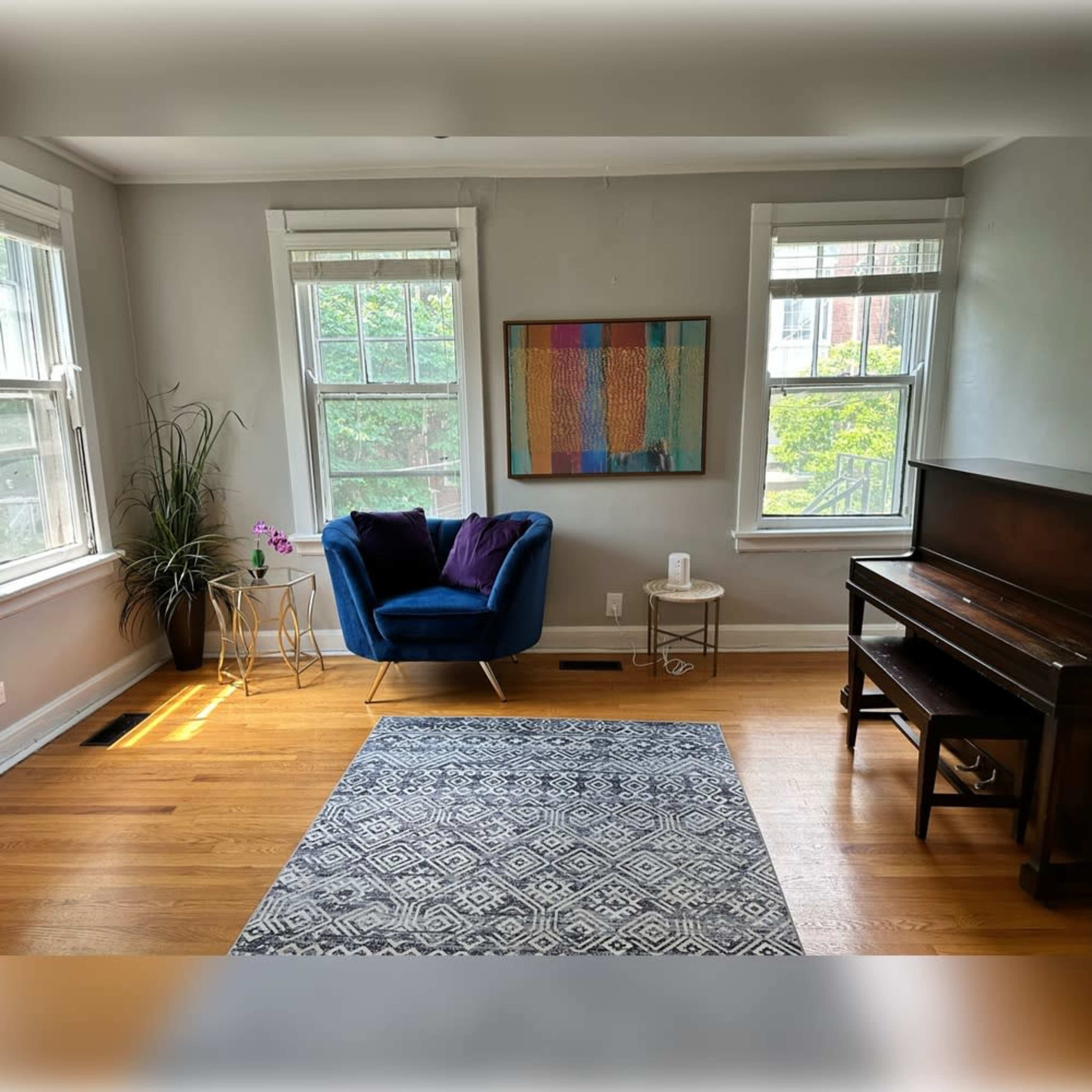 The image shows a bright living room featuring a blue armchair, a small side table, a piano, a patterned rug, and two windows that allow natural light to enter.