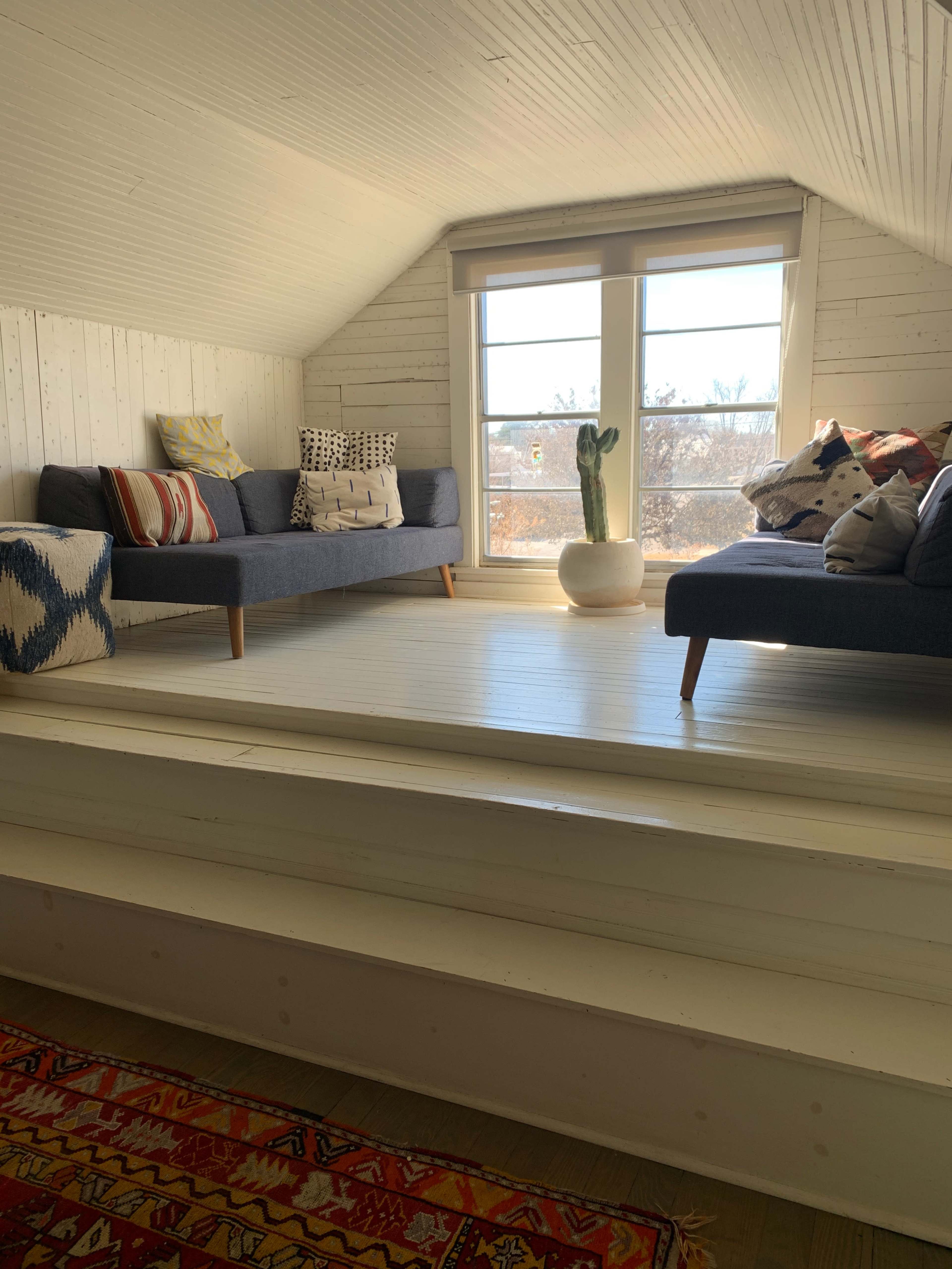 The image shows a bright attic space with two sofas facing large windows, a potted cactus, and a decorative rug on the floor.