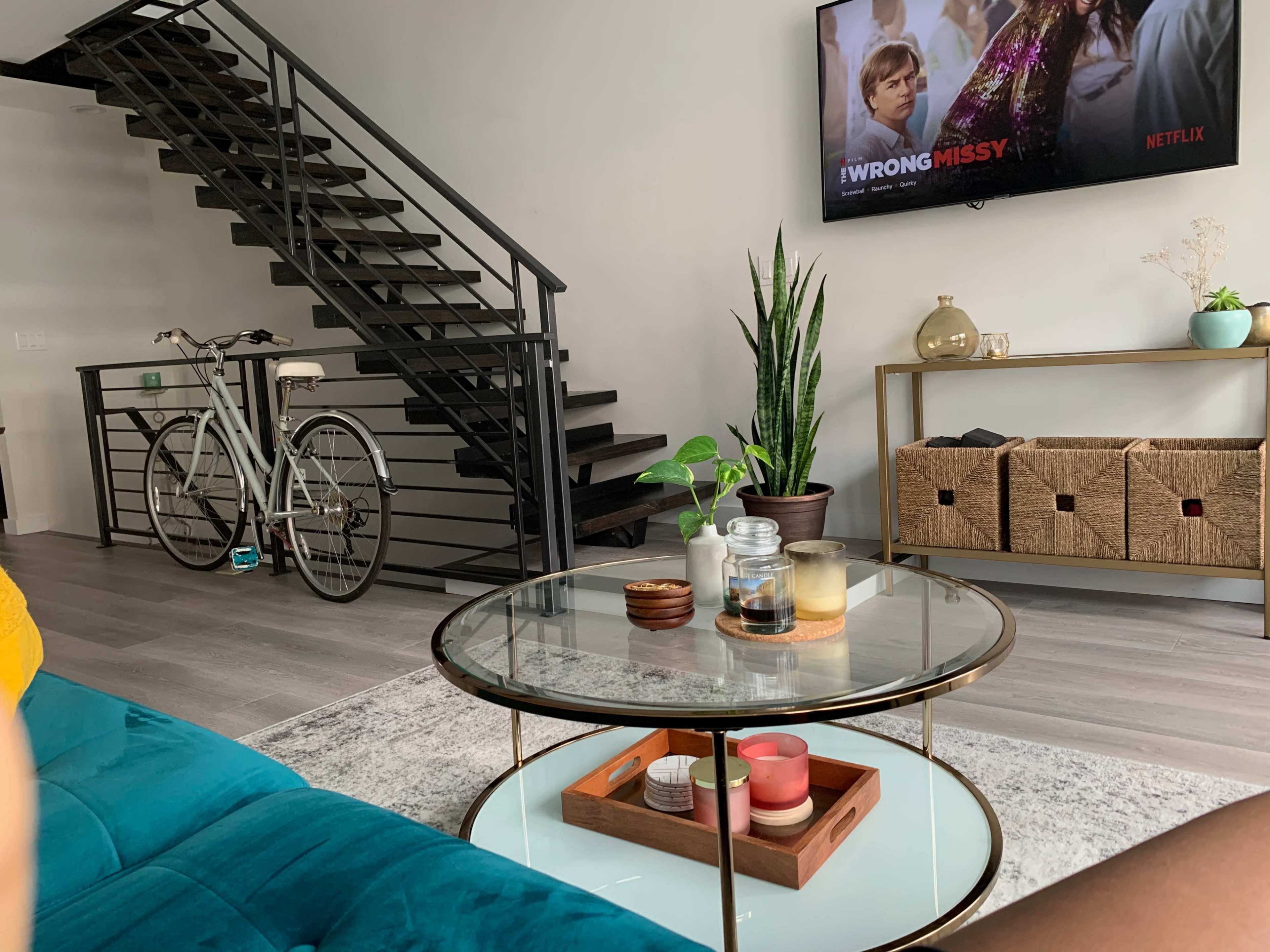A modern living room with a teal sofa, a glass coffee table, a staircase in the background, a bicycle, and a television displaying a Netflix show.