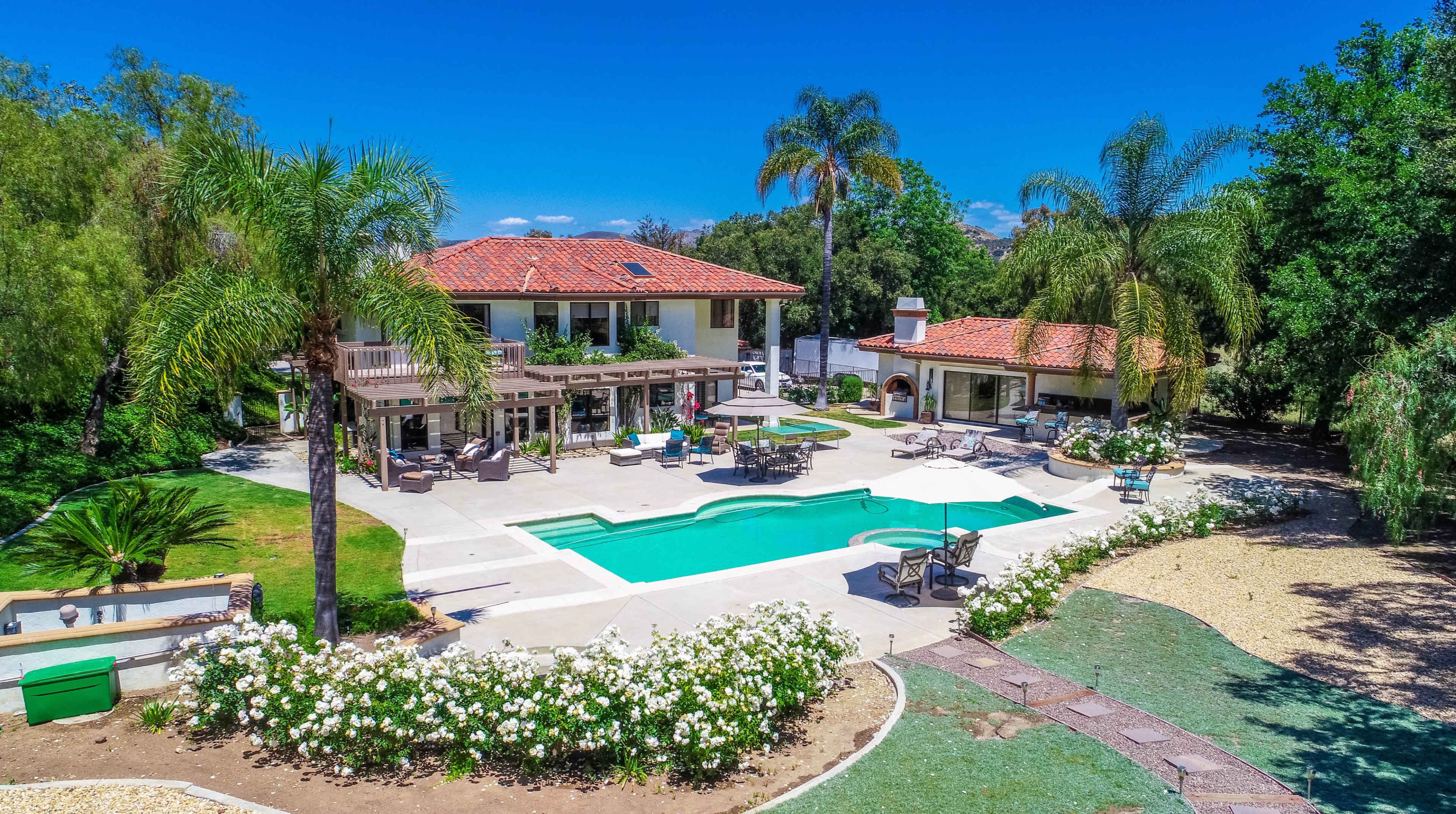 A large property featuring a pool surrounded by a landscaped yard, with a two-story house and an additional structure in the background.