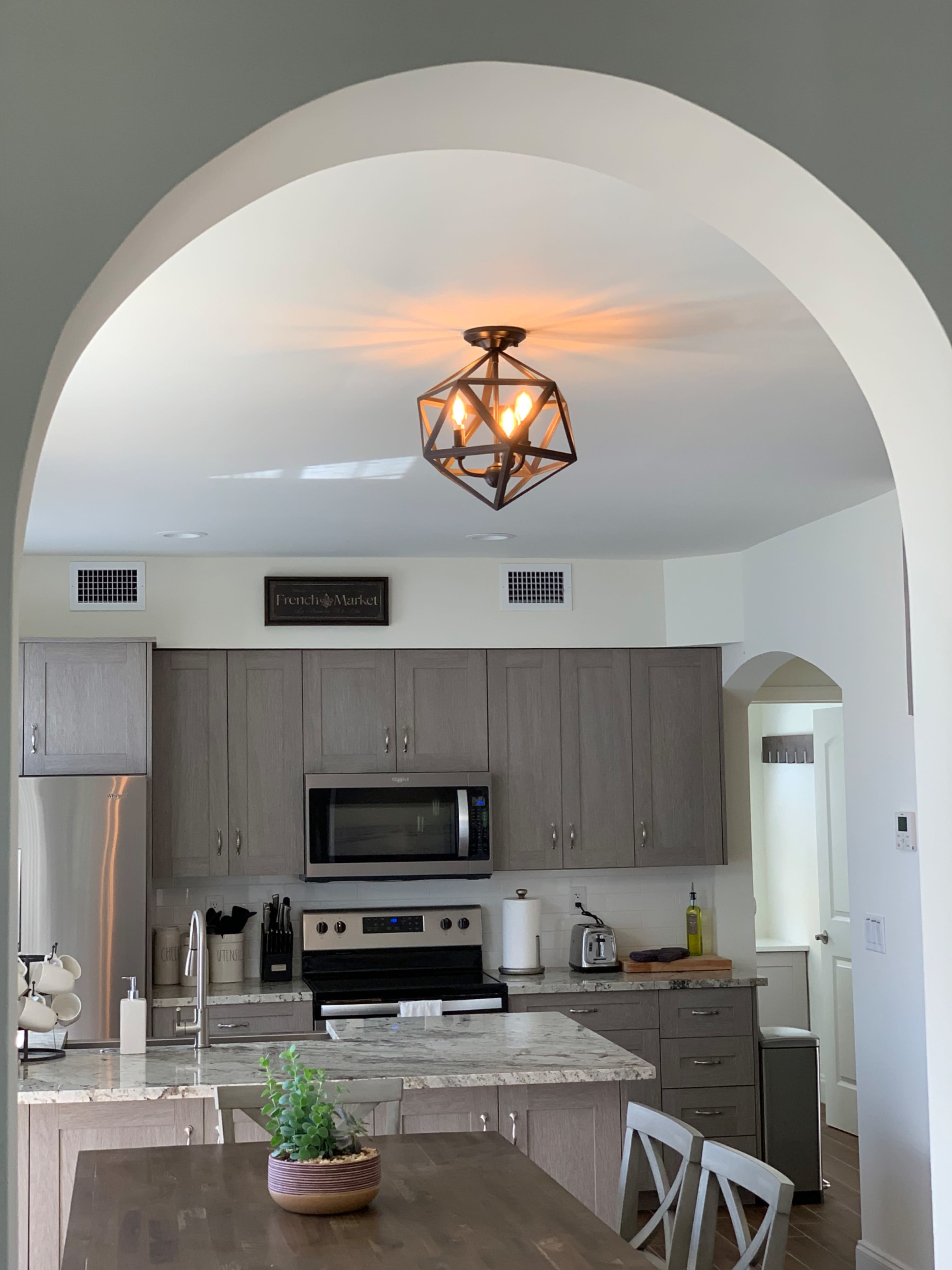 A kitchen features light wood cabinetry, stainless steel appliances, and a geometric pendant light hanging from an arched ceiling.