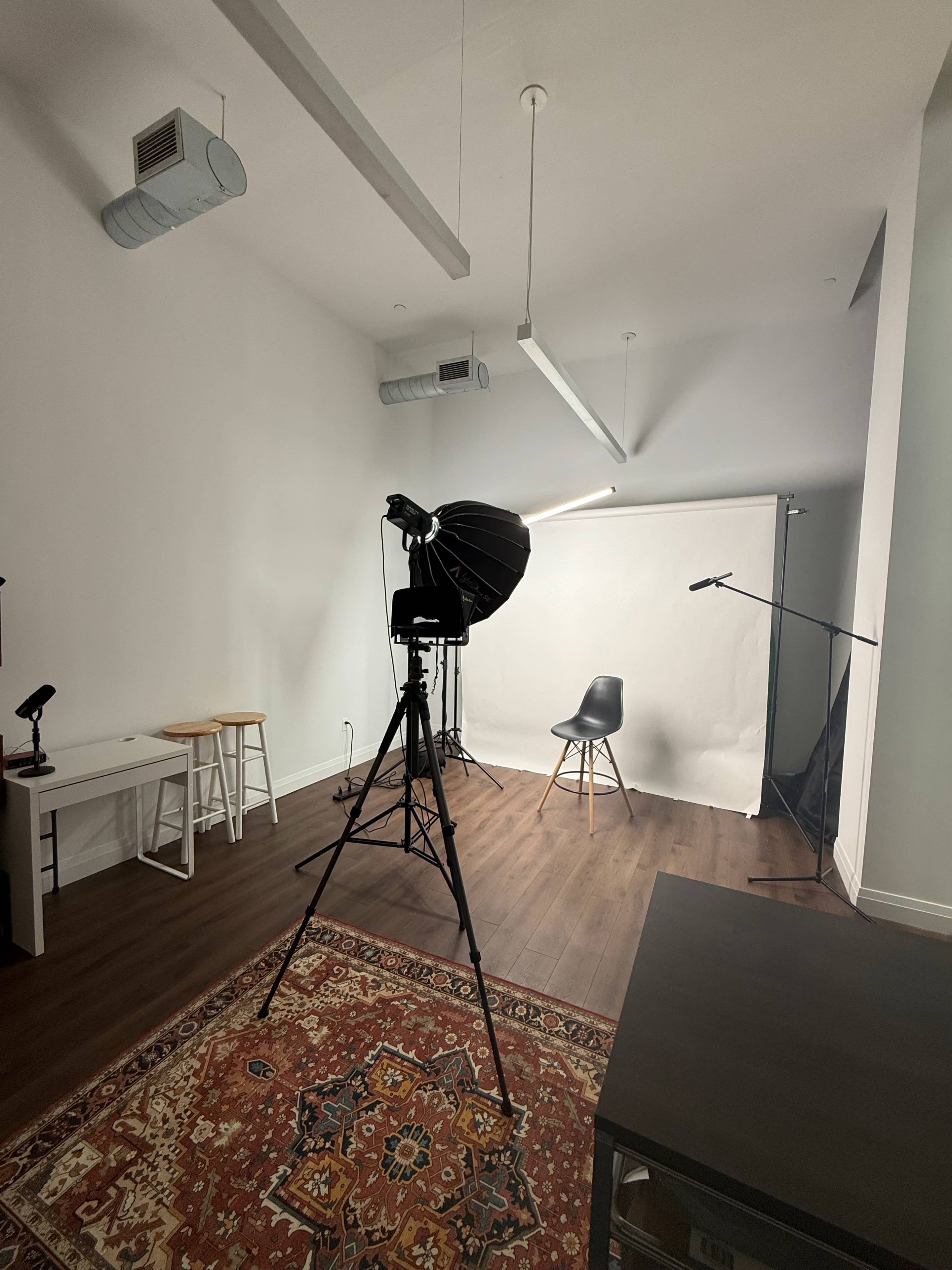 The image shows a photography studio with a large softbox light, a camera on a tripod, a black chair positioned in front of a backdrop, and a patterned rug on the floor.