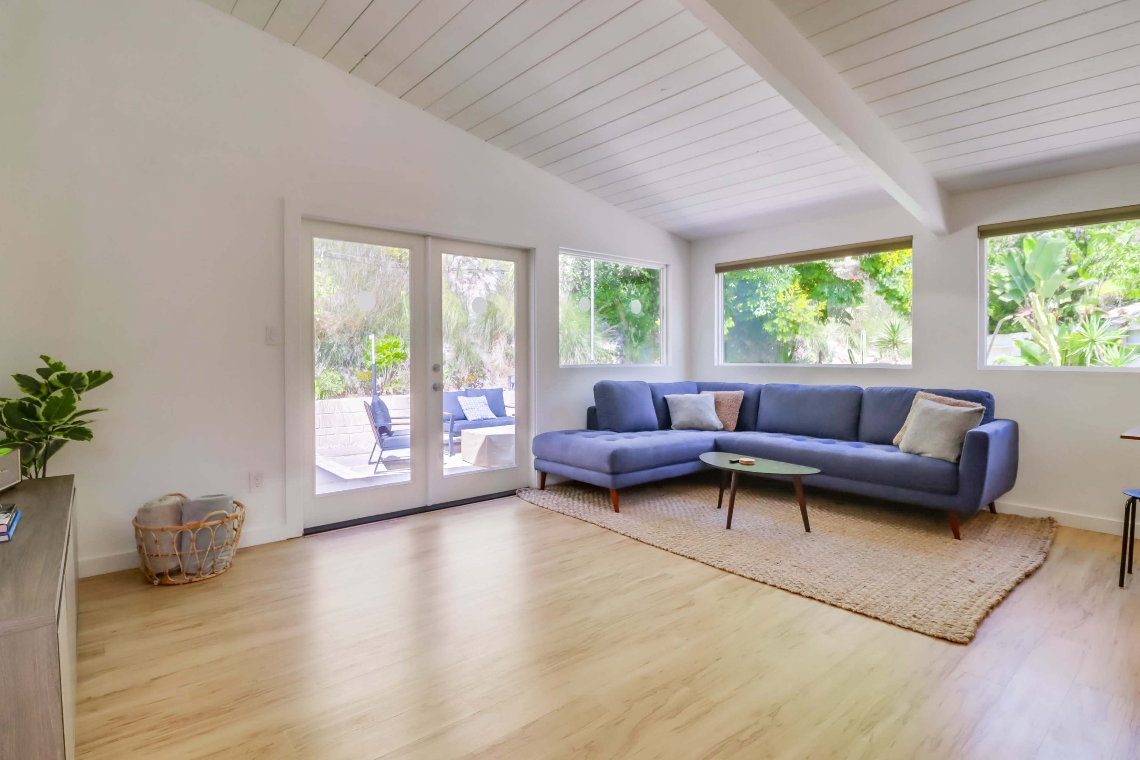 A modern living room features a blue sectional sofa, large windows allowing natural light, and a patio visible through sliding glass doors.