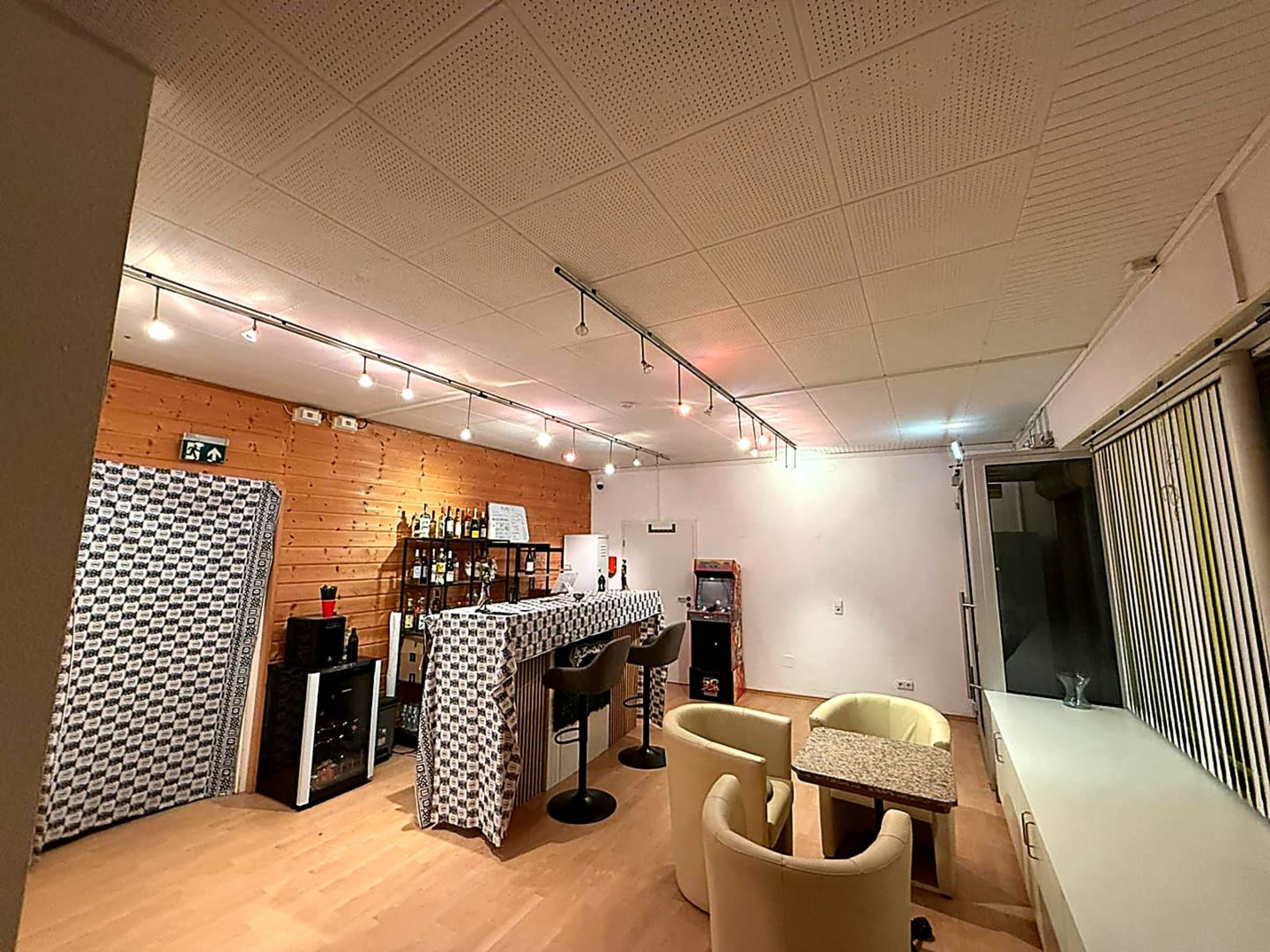 The image shows a room designed as a small bar, featuring a counter, a few chairs, and shelves with bottles against a wooden wall.