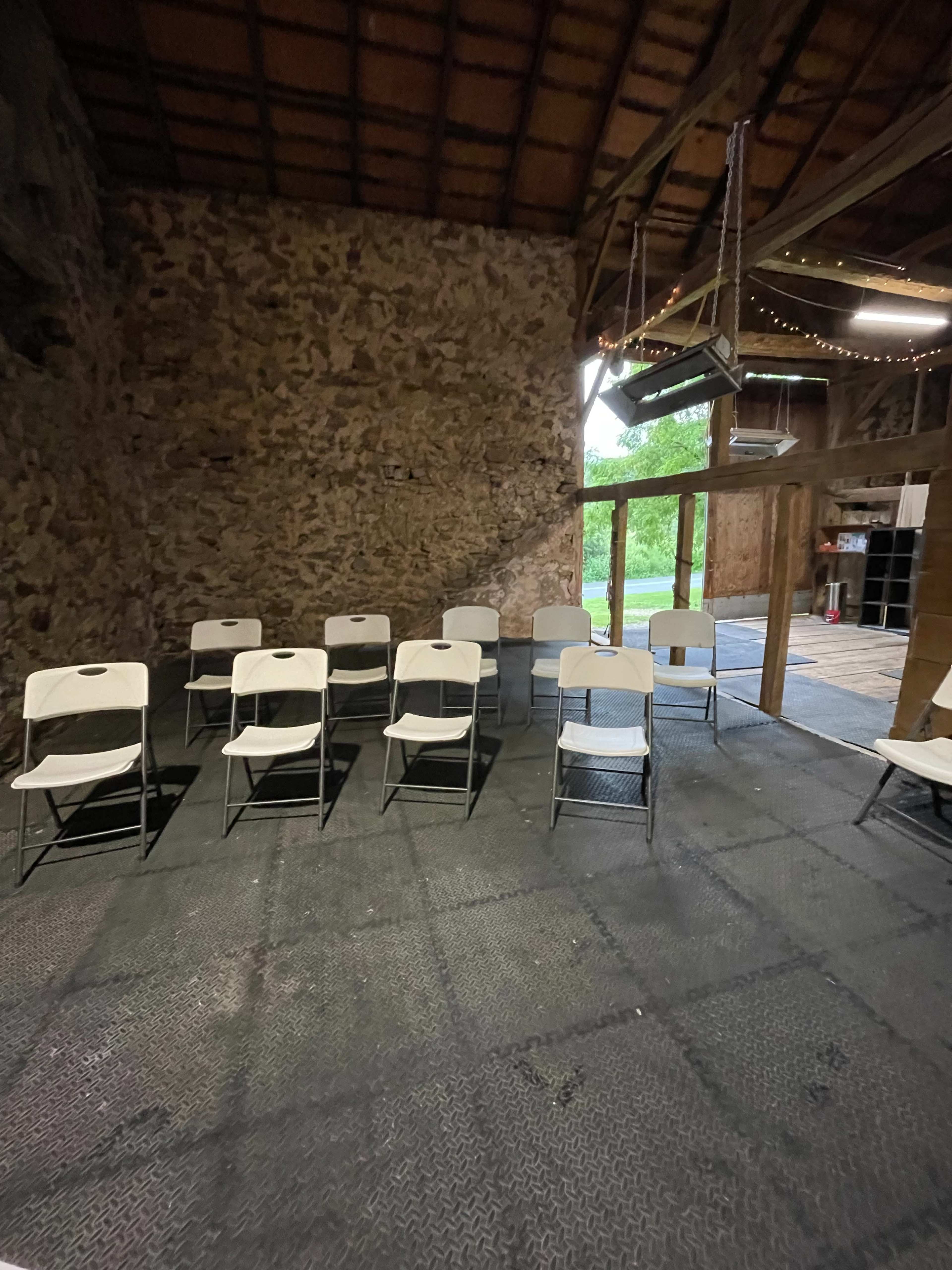 A row of white folding chairs is arranged on a textured black floor inside a rustic building with stone walls and wooden beams.