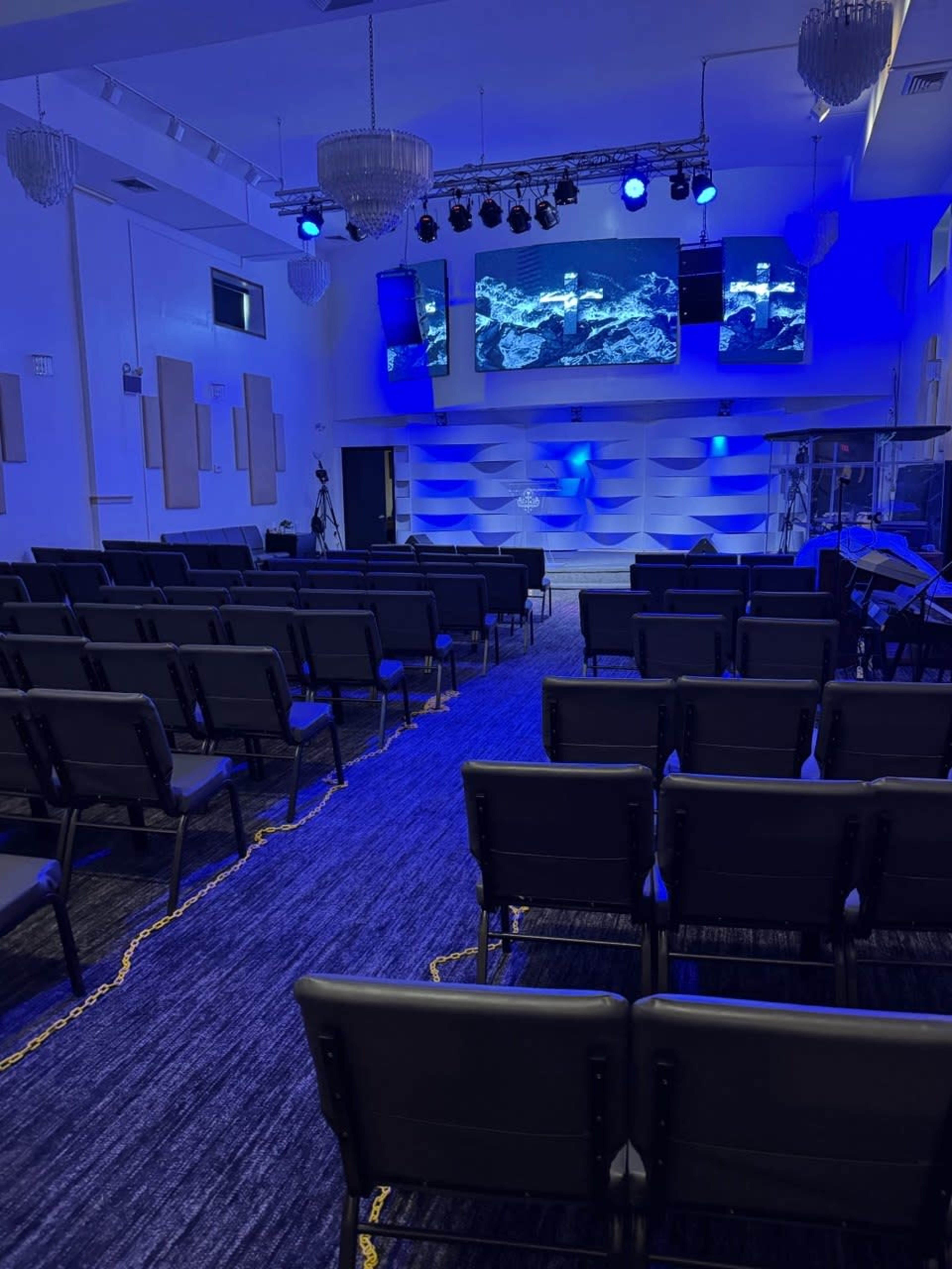 The image shows a modern, empty church interior with blue lighting, rows of black chairs, and large screens at the front displaying abstract visuals.