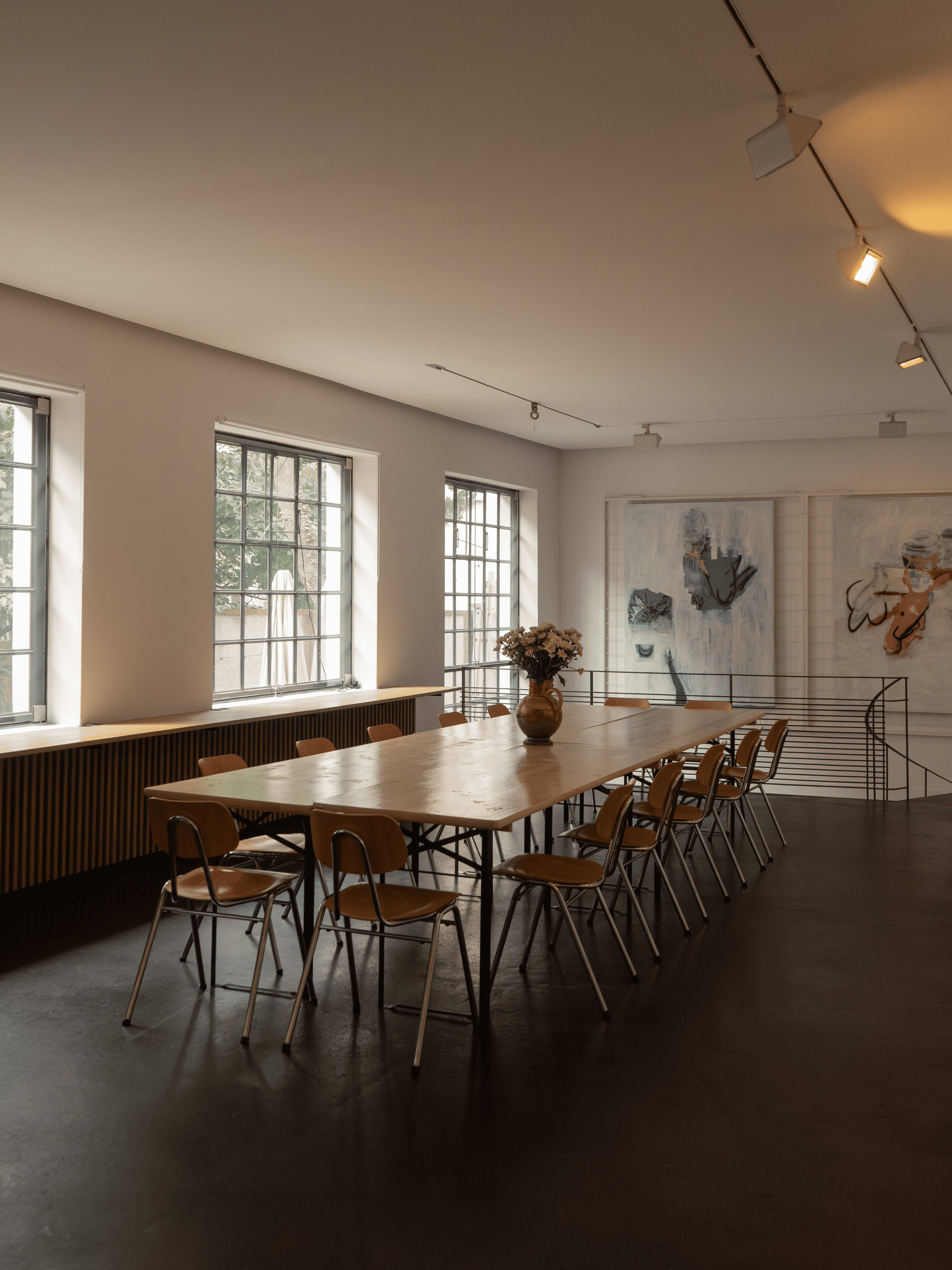 A spacious room features a long wooden table surrounded by metal chairs, with large windows and abstract artwork on the walls.