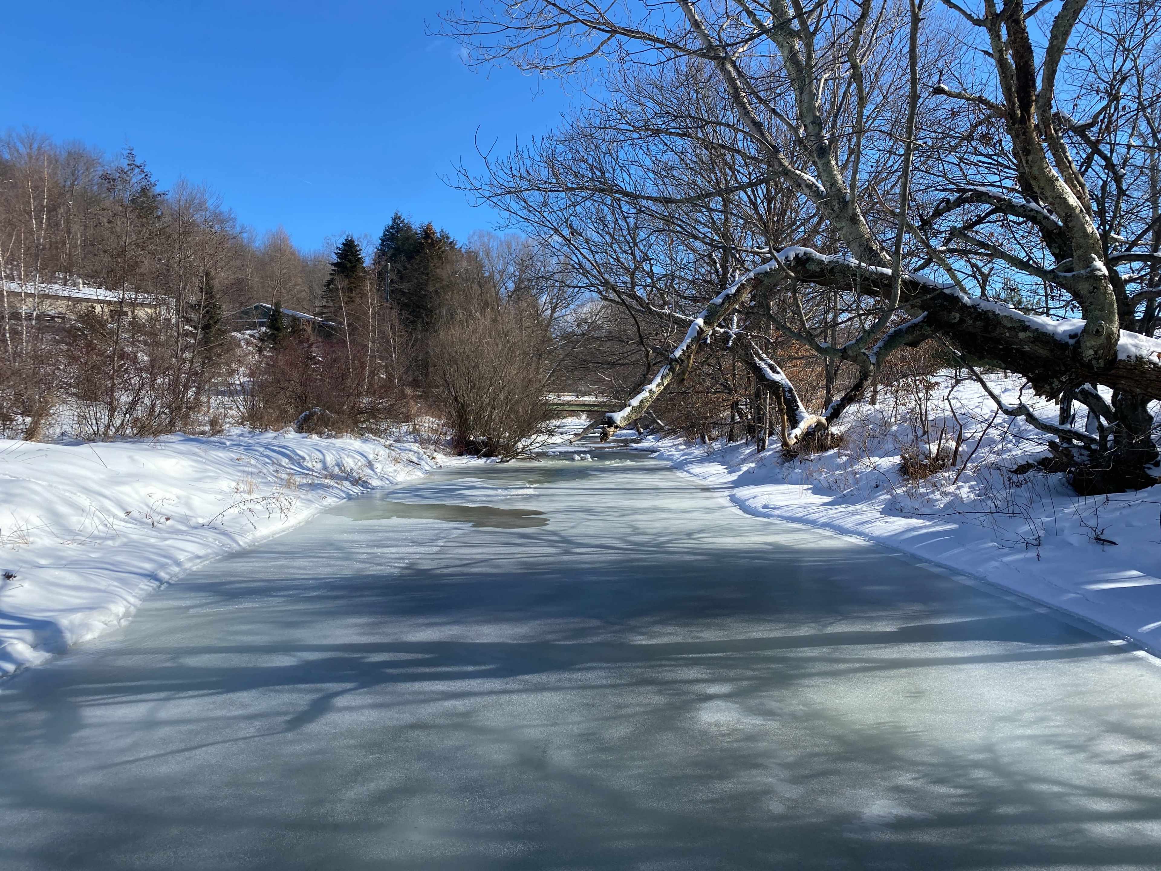A frozen river reflects the clear blue sky, bordered by snow-covered banks and leafless trees.