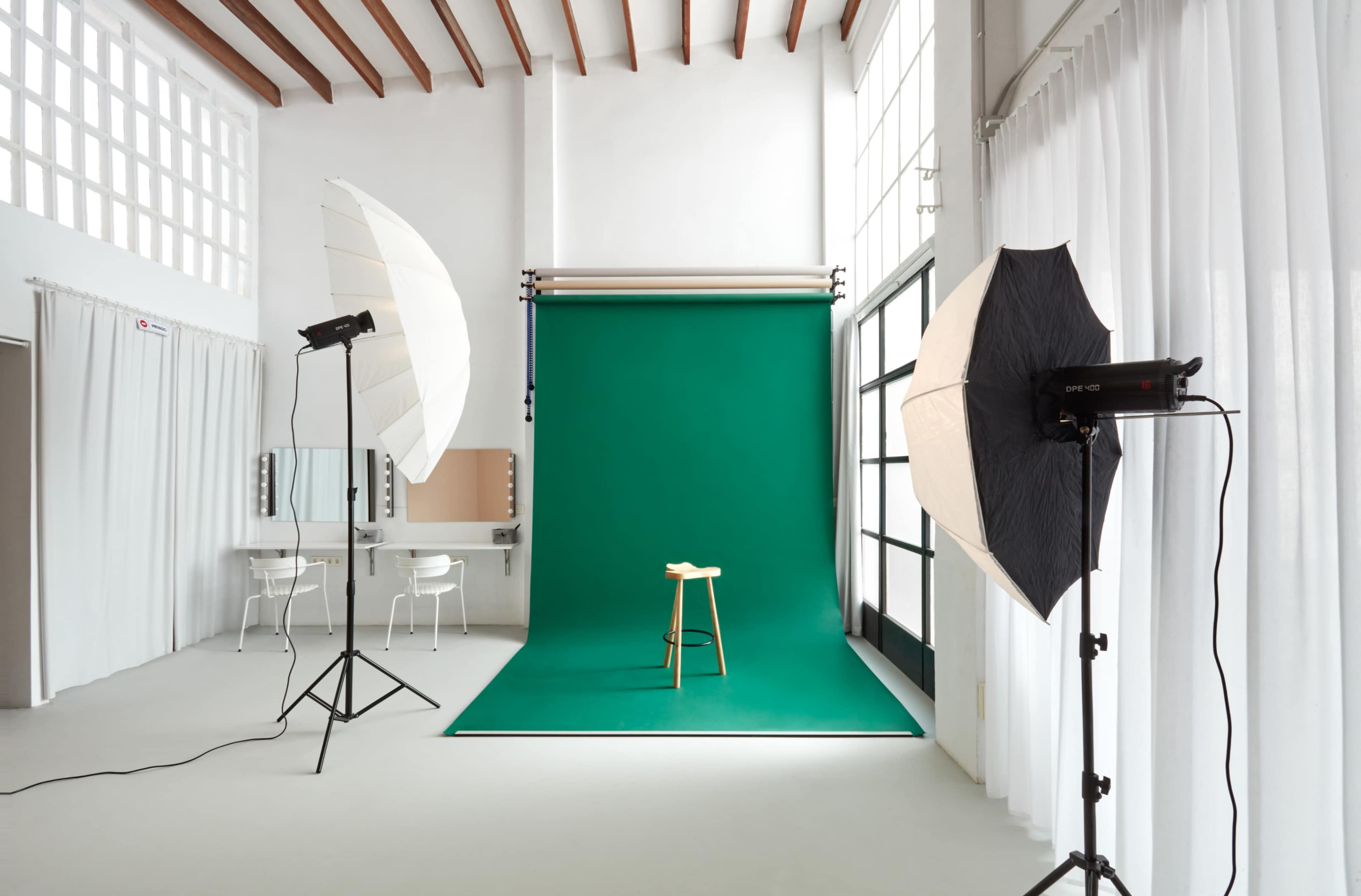 The image shows a photography studio with a green backdrop, a wooden stool placed in front, and studio lights positioned on either side.