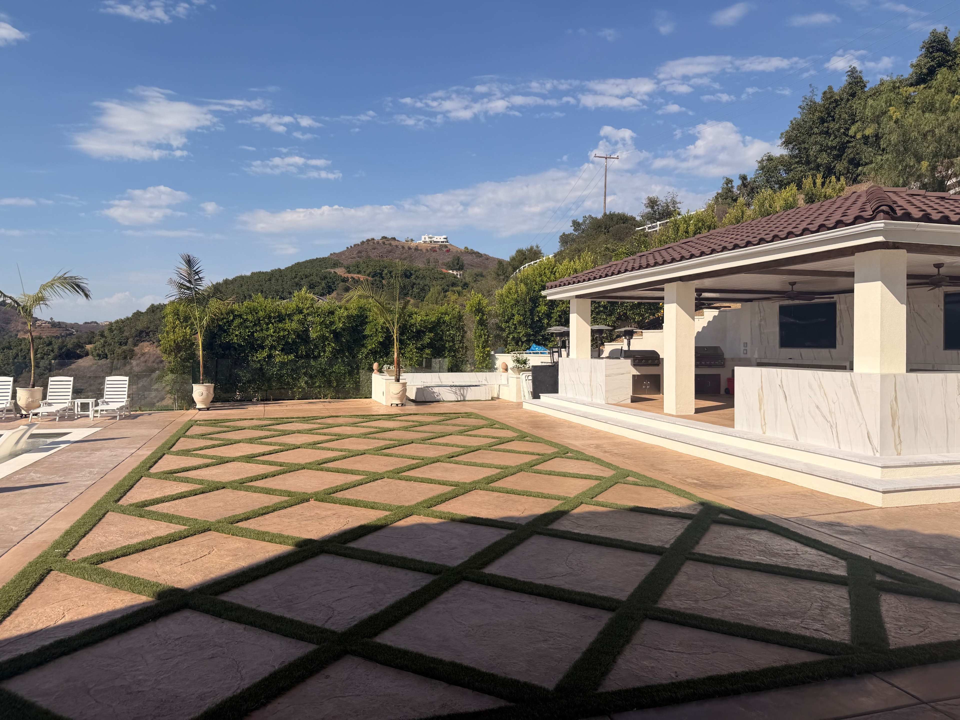 The image shows a spacious outdoor area with a patterned grass design, a nearby pool, and a covered outdoor kitchen or patio against a backdrop of hills.