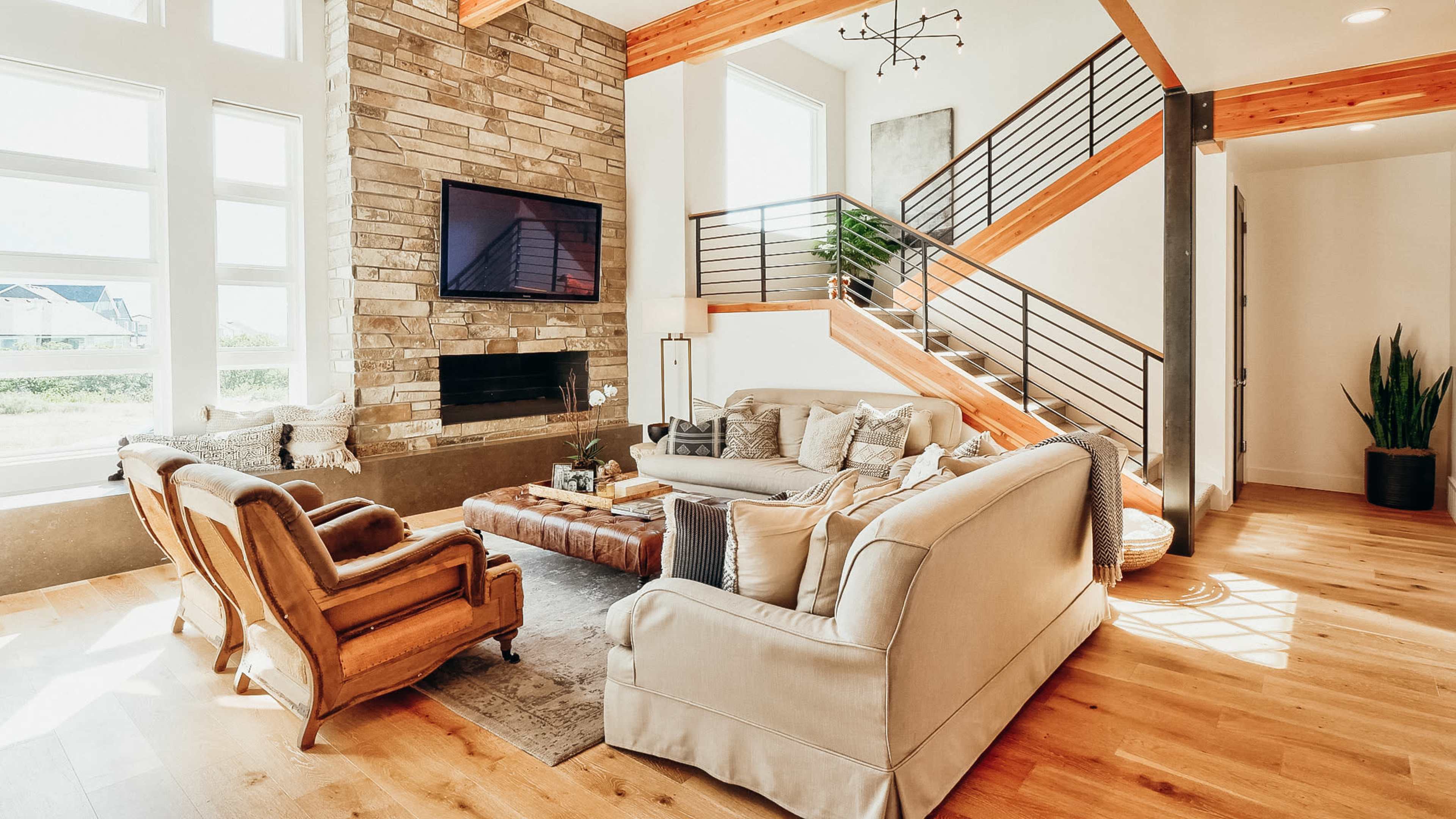 A modern living room features a stone fireplace, a sectional sofa, and a stylish staircase with wooden beams.