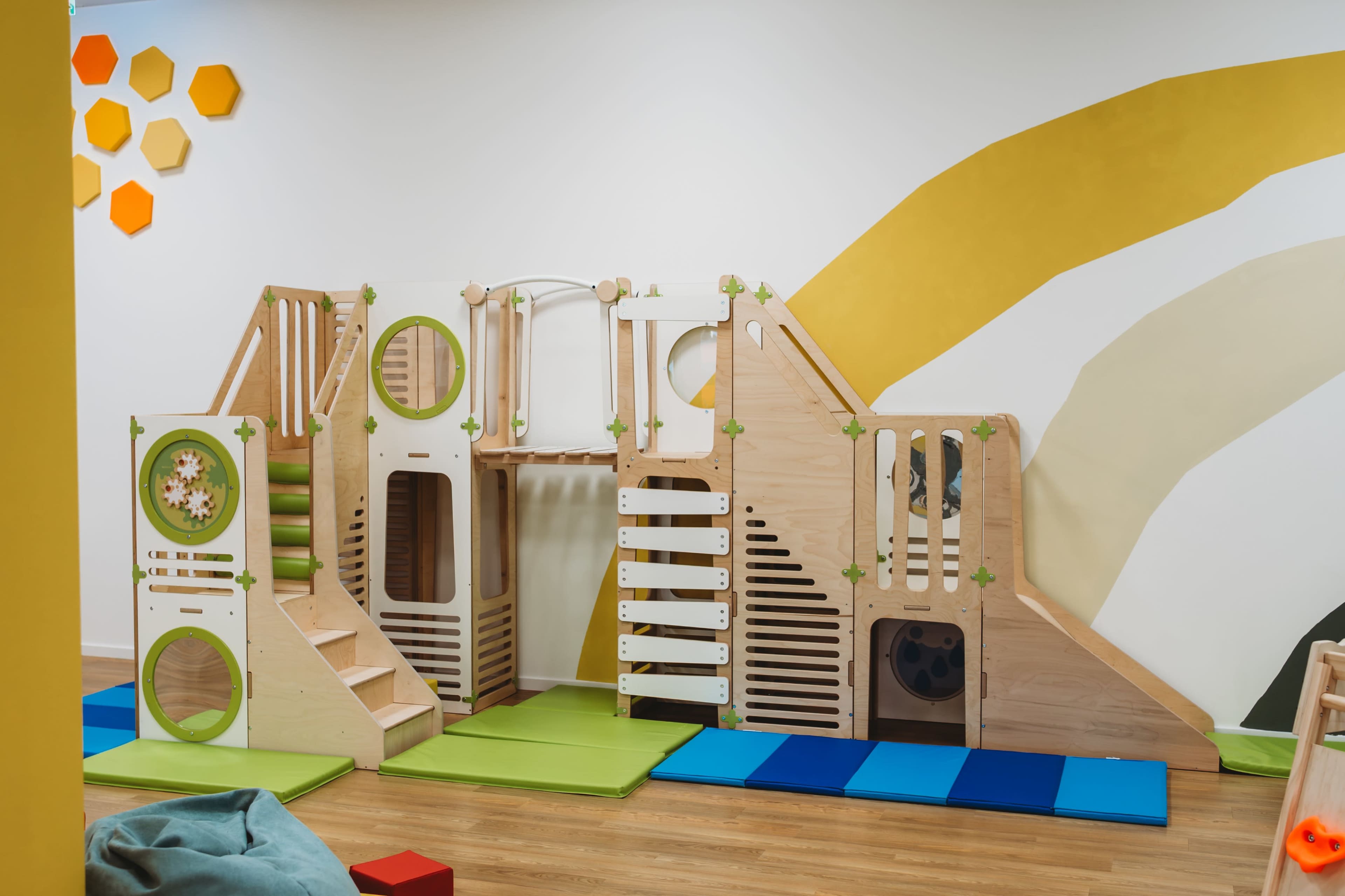 The image features a wooden play structure with climbing elements, slides, and various textures, set against a brightly colored wall in a playroom.