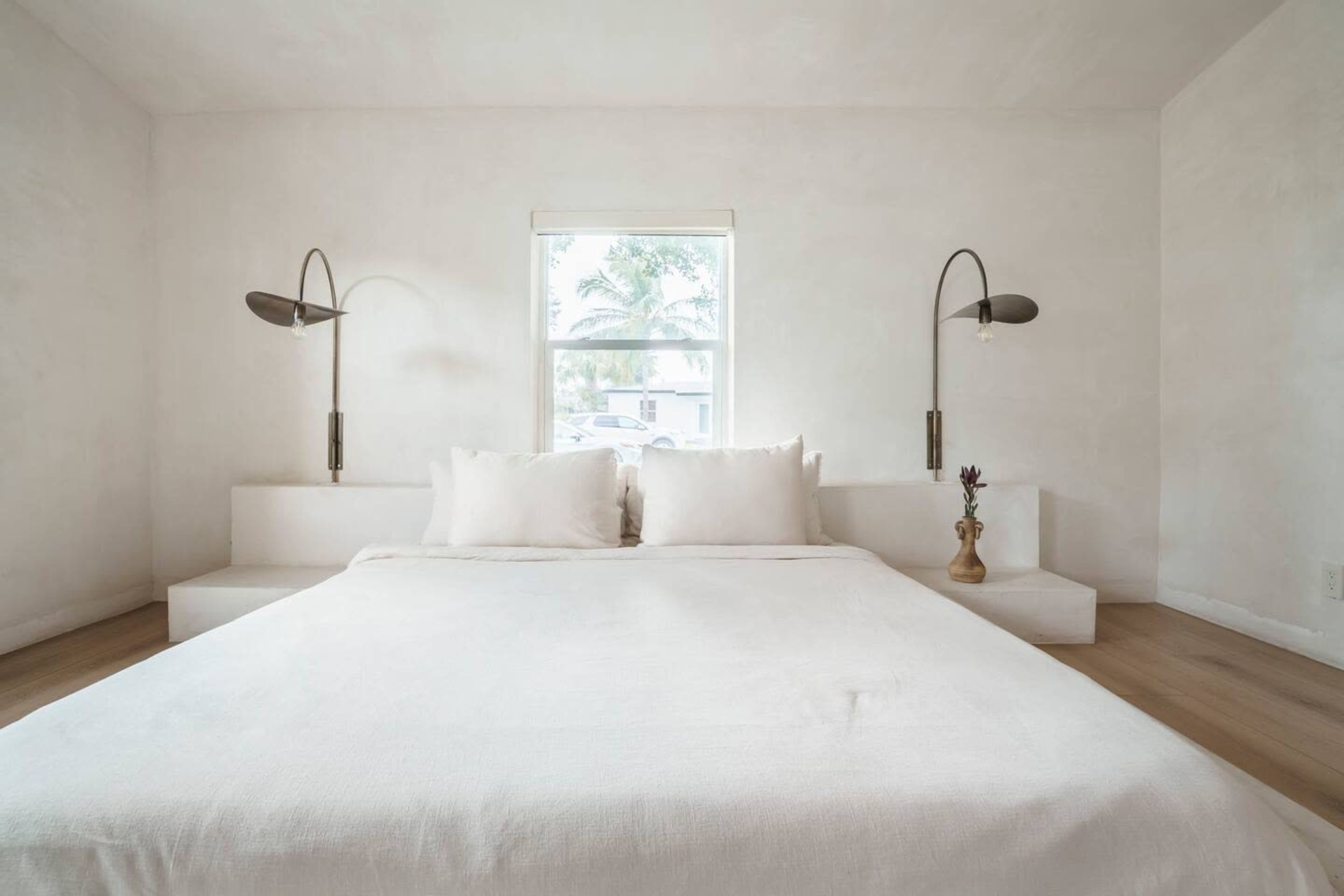 A minimalistic bedroom features a white bed with two large pillows, flanked by wall-mounted lamps and a window overlooking palm trees.