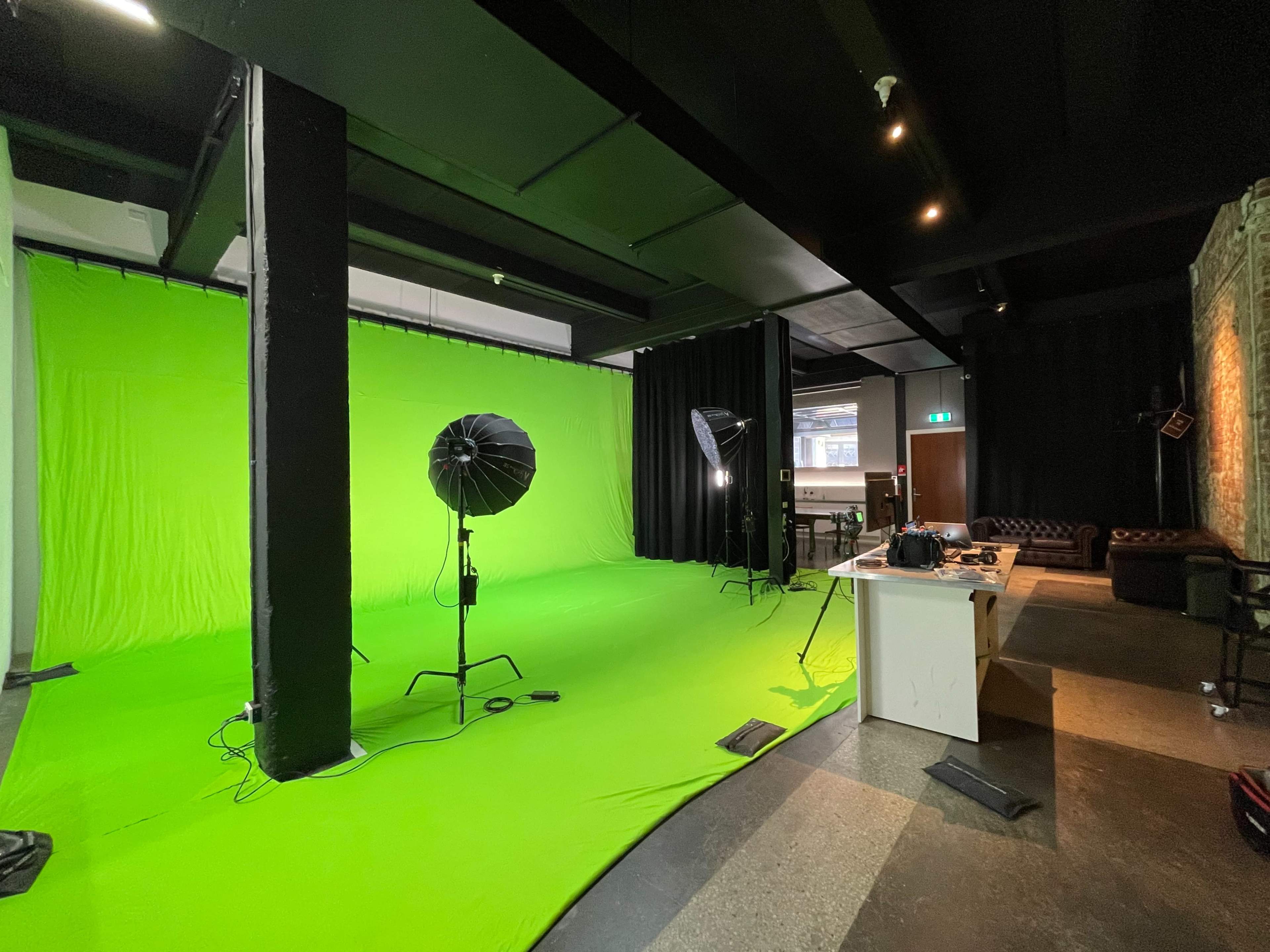 The image shows a photo studio featuring a large green screen, two softbox lights, and a preparation area with a couch and various equipment.