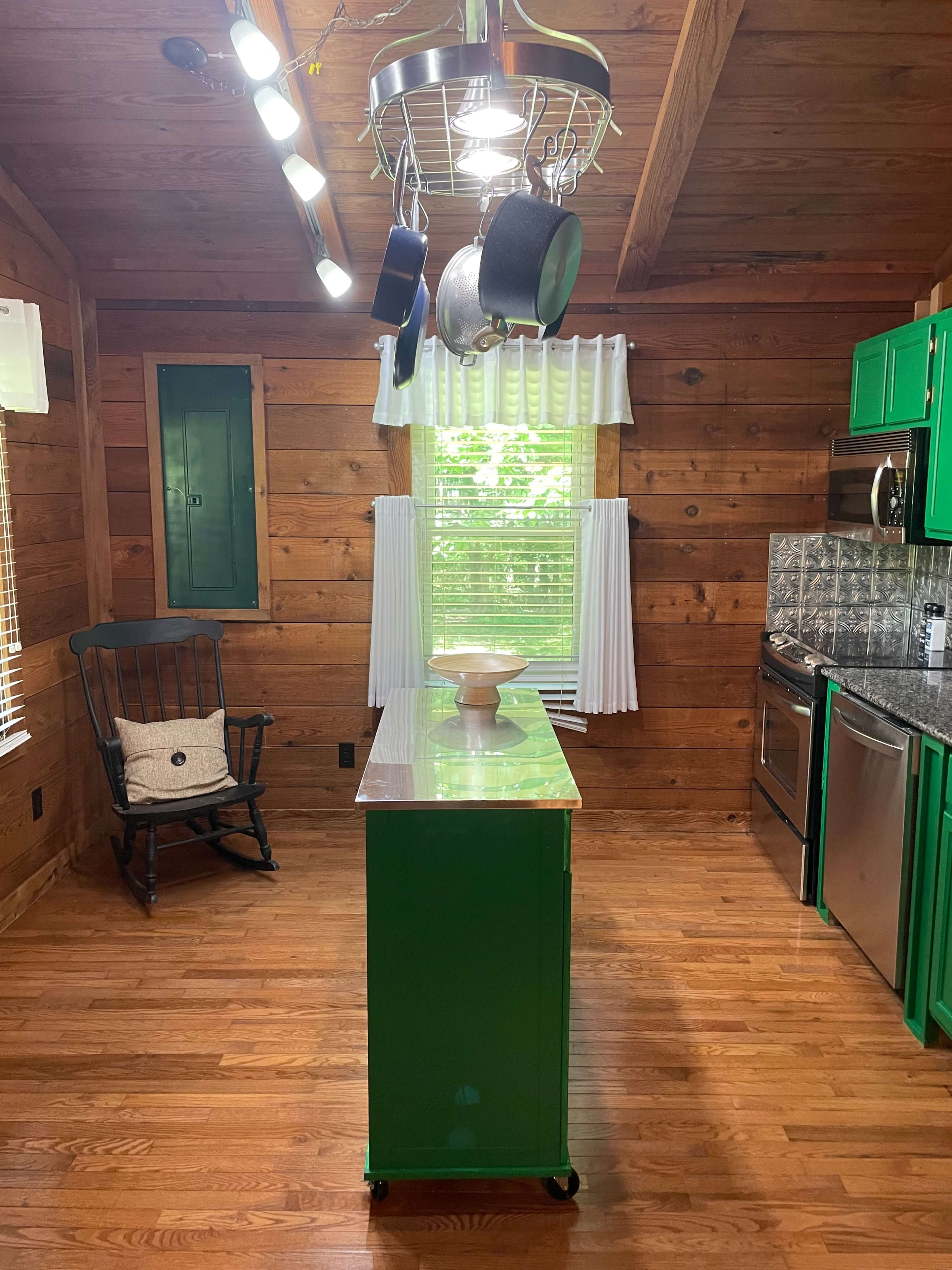 The image shows a wooden kitchen with green cabinets, a pot rack hanging from the ceiling, and a central island with a wooden countertop.
